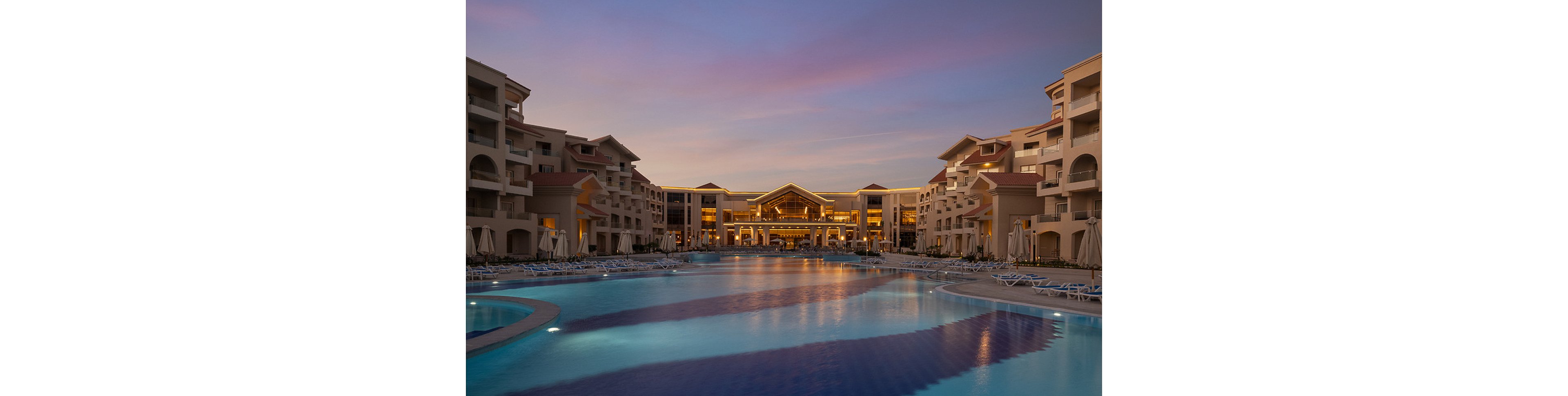 A grand resort complex is showcased at dusk, with its main building and wings warmly illuminated. The expansive swimming pool in the foreground reflects the soft purple and blue hues of the evening sky. Lounge chairs and umbrellas line the poolside, suggesting a tranquil and upscale vacation setting. The architecture features multiple balconies and a symmetrical design, enhancing the sense of luxury and relaxation.