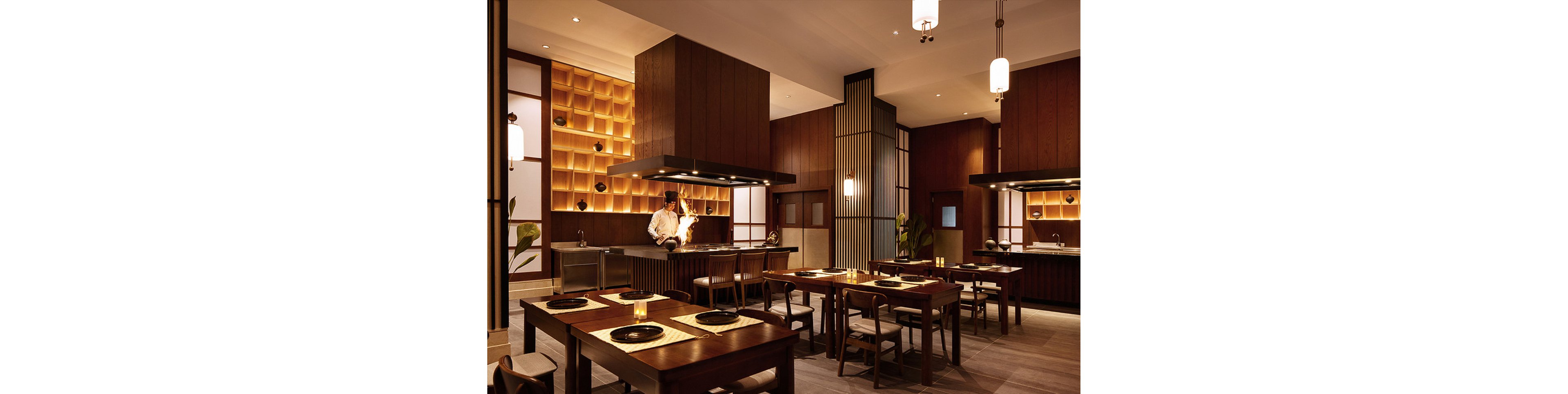 A contemporary Japanese restaurant features wooden tables and chairs, with a chef preparing food at an open kitchen counter. Warm lighting and minimalist decor create a cozy, inviting atmosphere. The setting is clean and organized, with decorative shelving and modern fixtures. No visible text or numbers are present in the image.