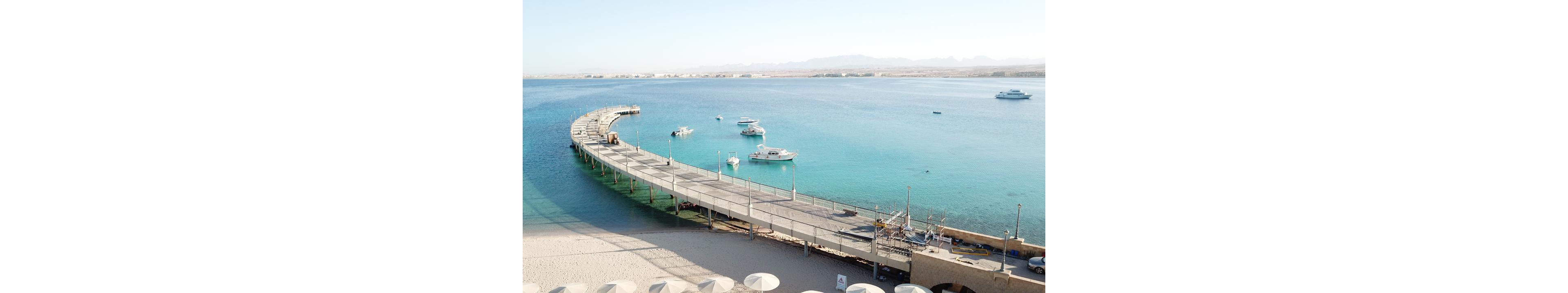 A long, curved pier stretches out over calm, turquoise waters with several small boats anchored nearby. The setting is a tranquil coastal area with a sandy beach in the foreground and distant mountains on the horizon. The scene is bright and serene, evoking a relaxed, vacation-like atmosphere. The water is clear, and the overall palette is dominated by blues and sandy neutrals.