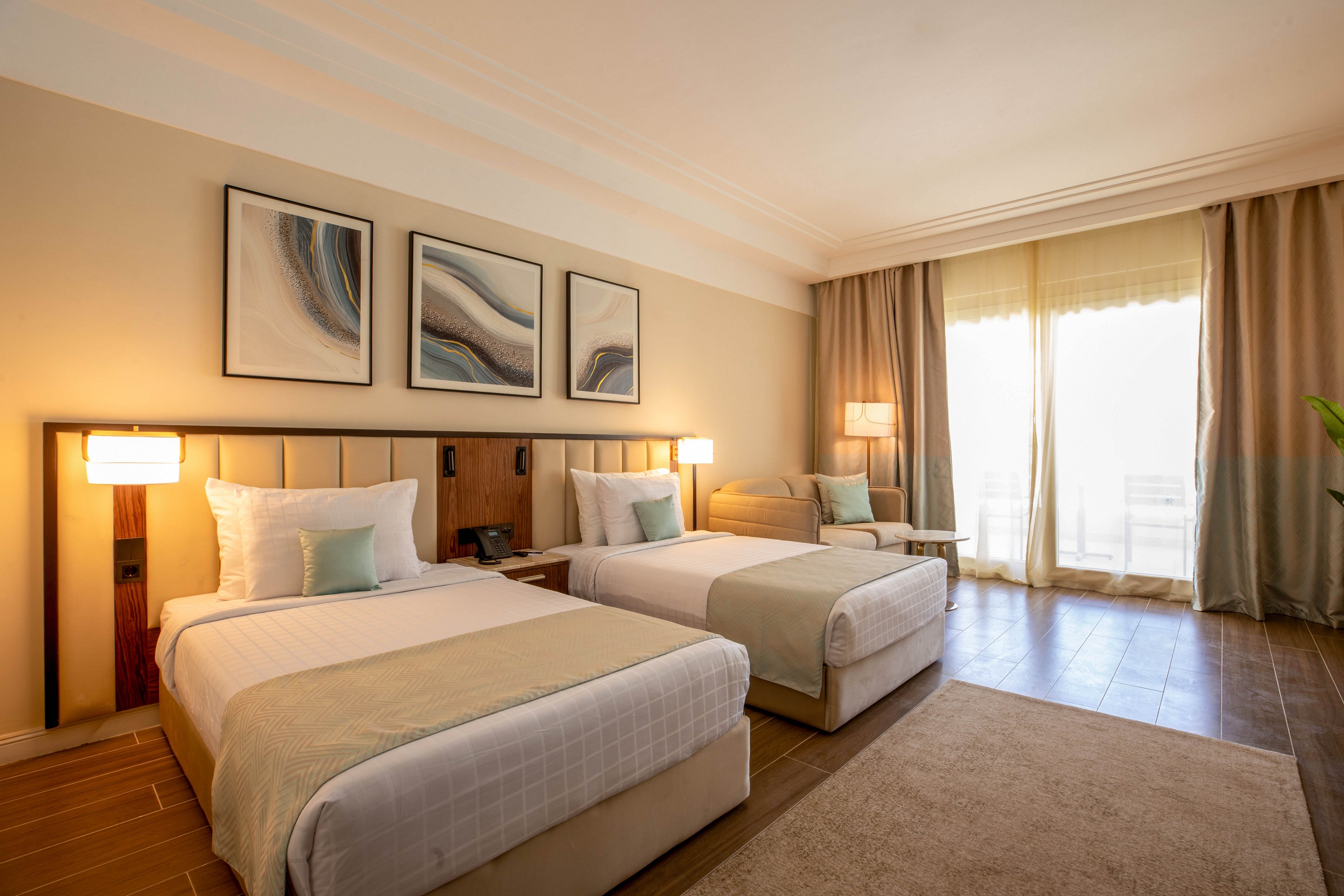 A contemporary hotel room features two neatly made twin beds with light bedding and accent pillows. The space is illuminated by natural light streaming through a large window with sheer curtains, complemented by warm bedside lamps. Abstract artwork decorates the wall above the beds, and a cozy seating area is visible in the corner. The room has a neutral color palette with beige and soft blue tones, creating a calm and inviting atmosphere.