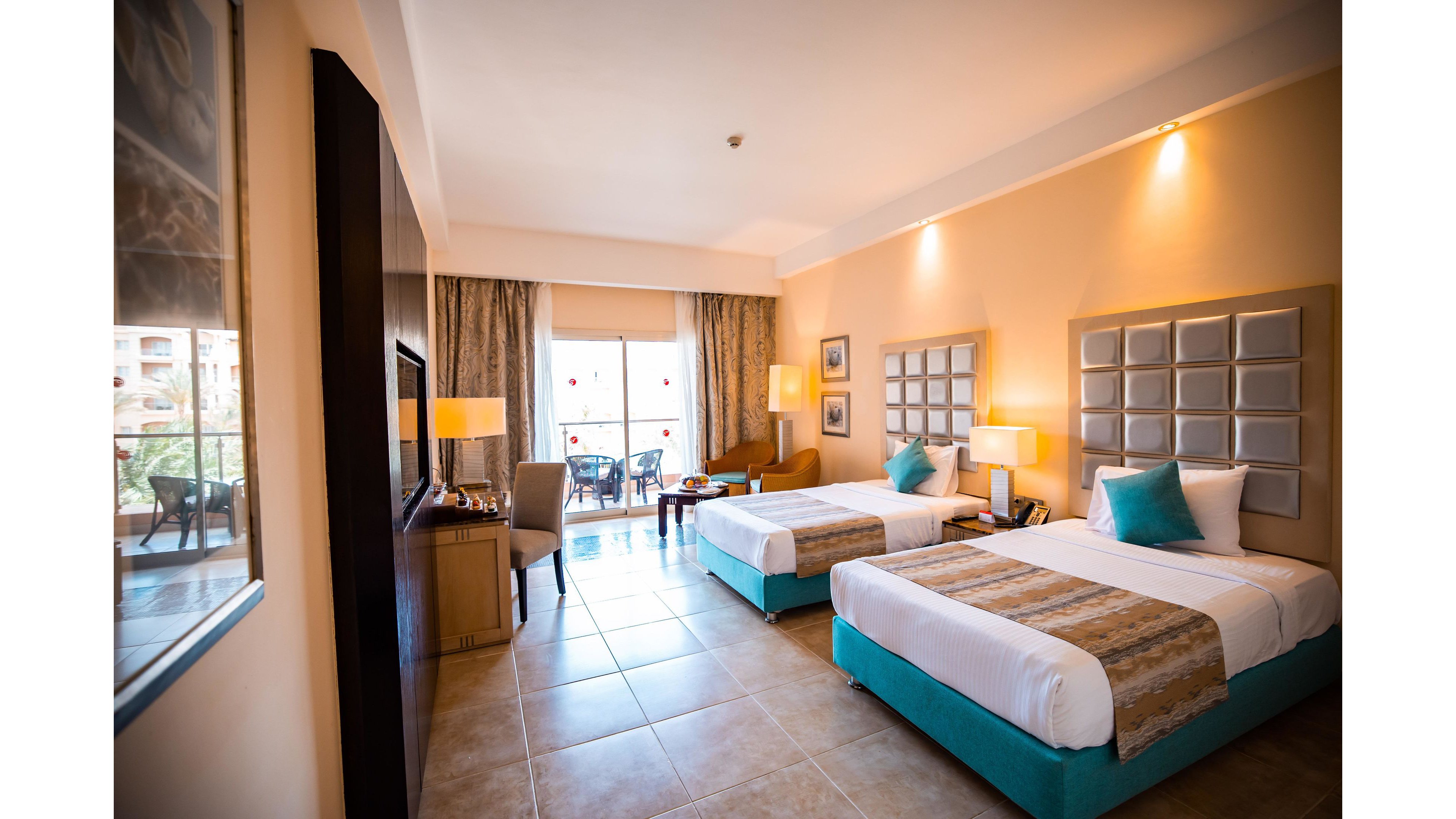 A spacious hotel room features two neatly made twin beds with turquoise accents and padded headboards. The room is well-lit with warm lighting, tiled floors, and a large window leading to a balcony with outdoor seating. Neutral tones and contemporary decor create a welcoming, relaxed atmosphere. No visible text or numbers are present in the image.