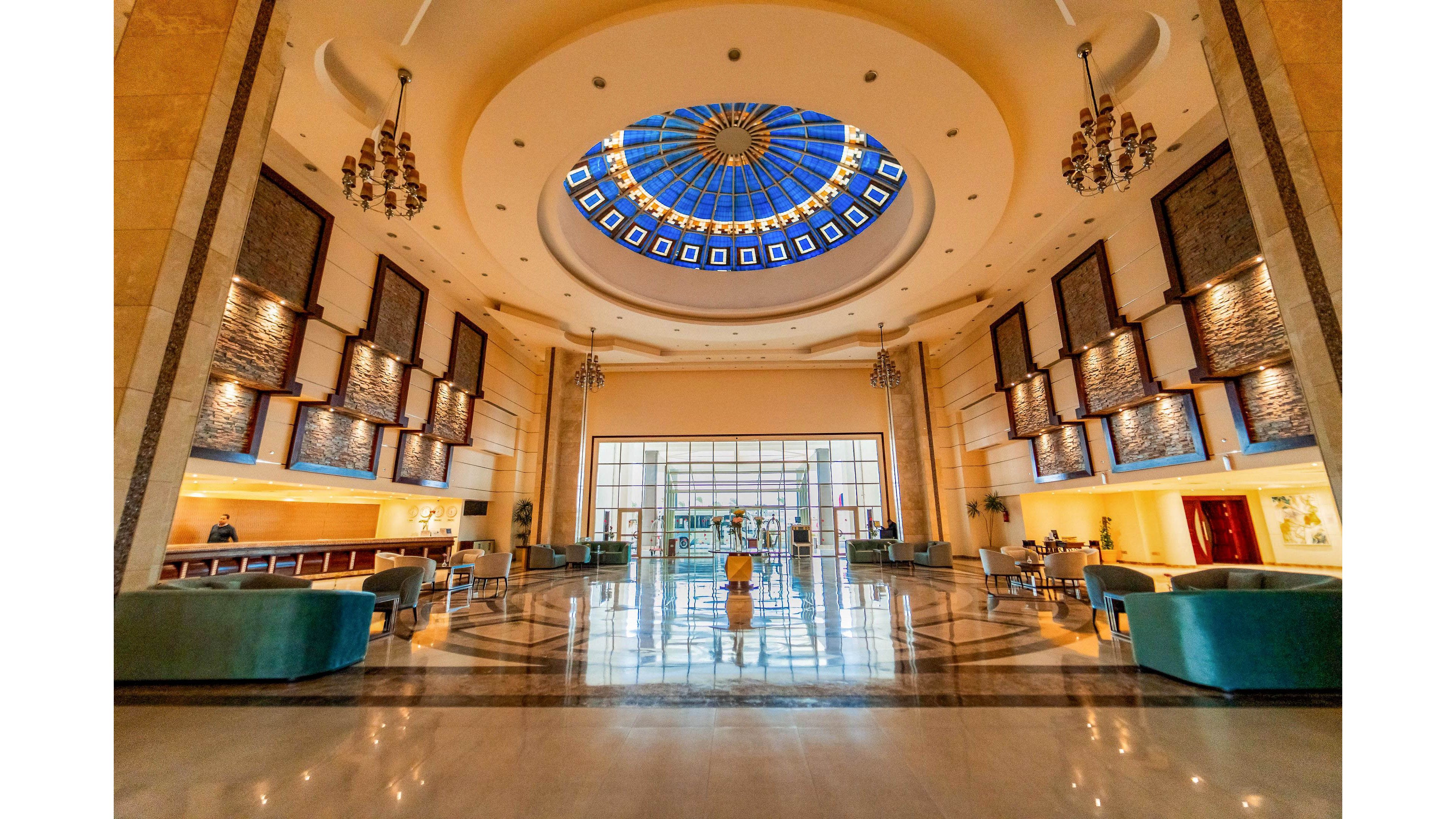 A spacious and luxurious hotel lobby features a striking circular glass dome ceiling with blue accents. The setting includes elegant chandeliers, marble floors, and plush seating areas, creating a welcoming and upscale atmosphere. Natural light floods the space through large windows at the far end, highlighting the modern architectural details. The overall mood is sophisticated and inviting, ideal for hospitality environments.