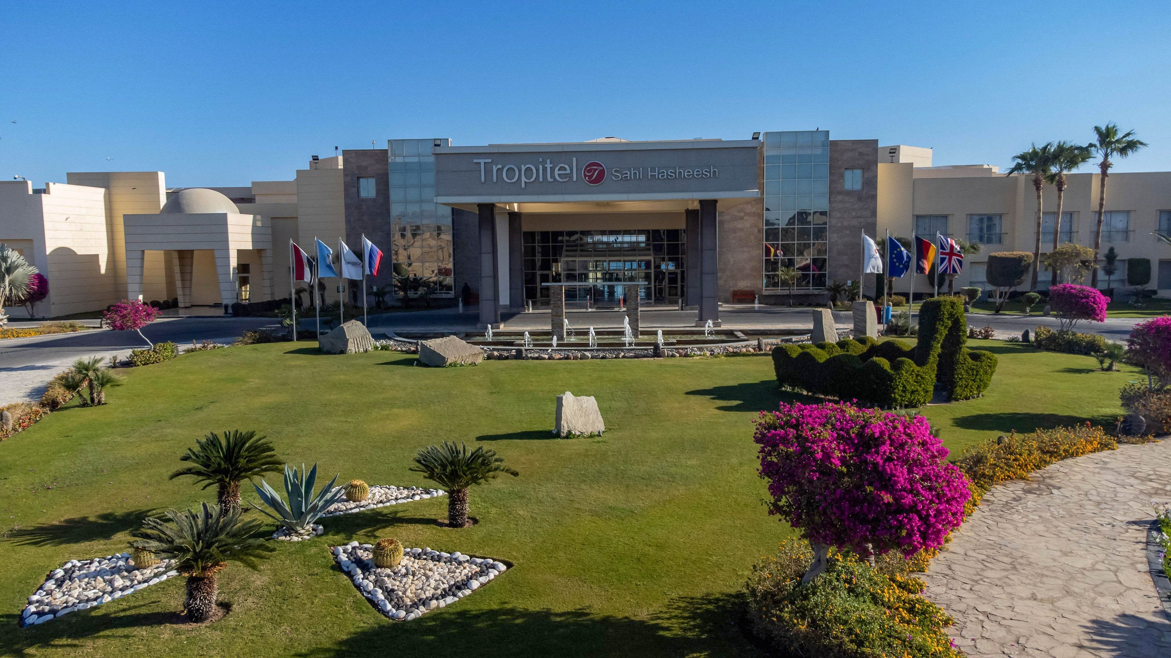 The image shows the main entrance of the Tropitel hotel, clearly identified by the signage above the entrance. The setting features a well-maintained garden with trimmed bushes, palm trees, and vibrant flowering plants. Multiple international flags are displayed near the entrance, adding a welcoming and global atmosphere. The sky is clear and blue, contributing to a bright and inviting mood. No people are visible in the image.
