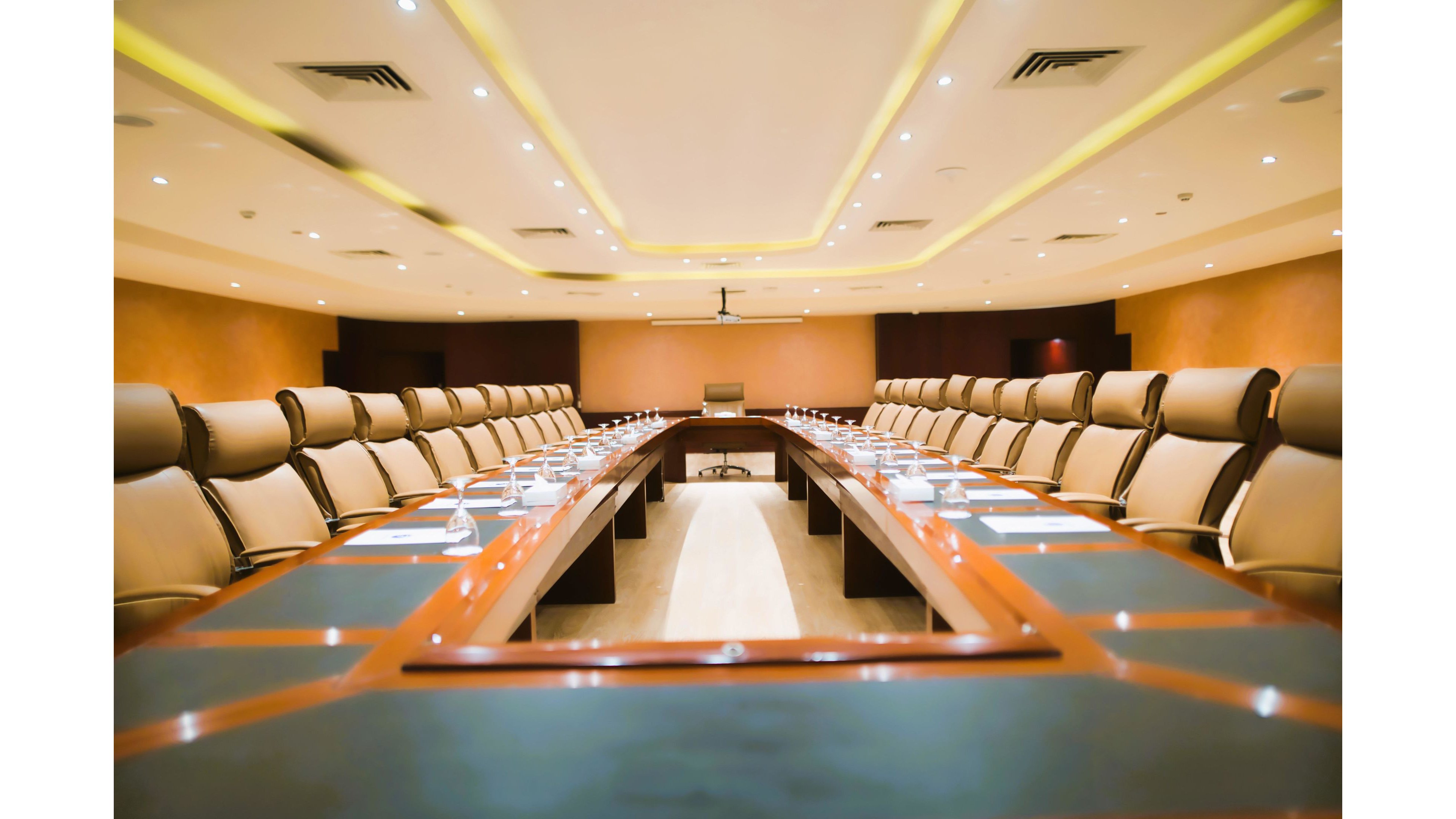 A spacious, well-lit boardroom features a long conference table surrounded by plush leather chairs. The room is empty, with water glasses and microphones neatly arranged at each seat, suggesting readiness for a formal meeting. Warm lighting and neutral tones create a professional and inviting atmosphere. The ceiling has recessed lighting and the overall style is contemporary corporate.