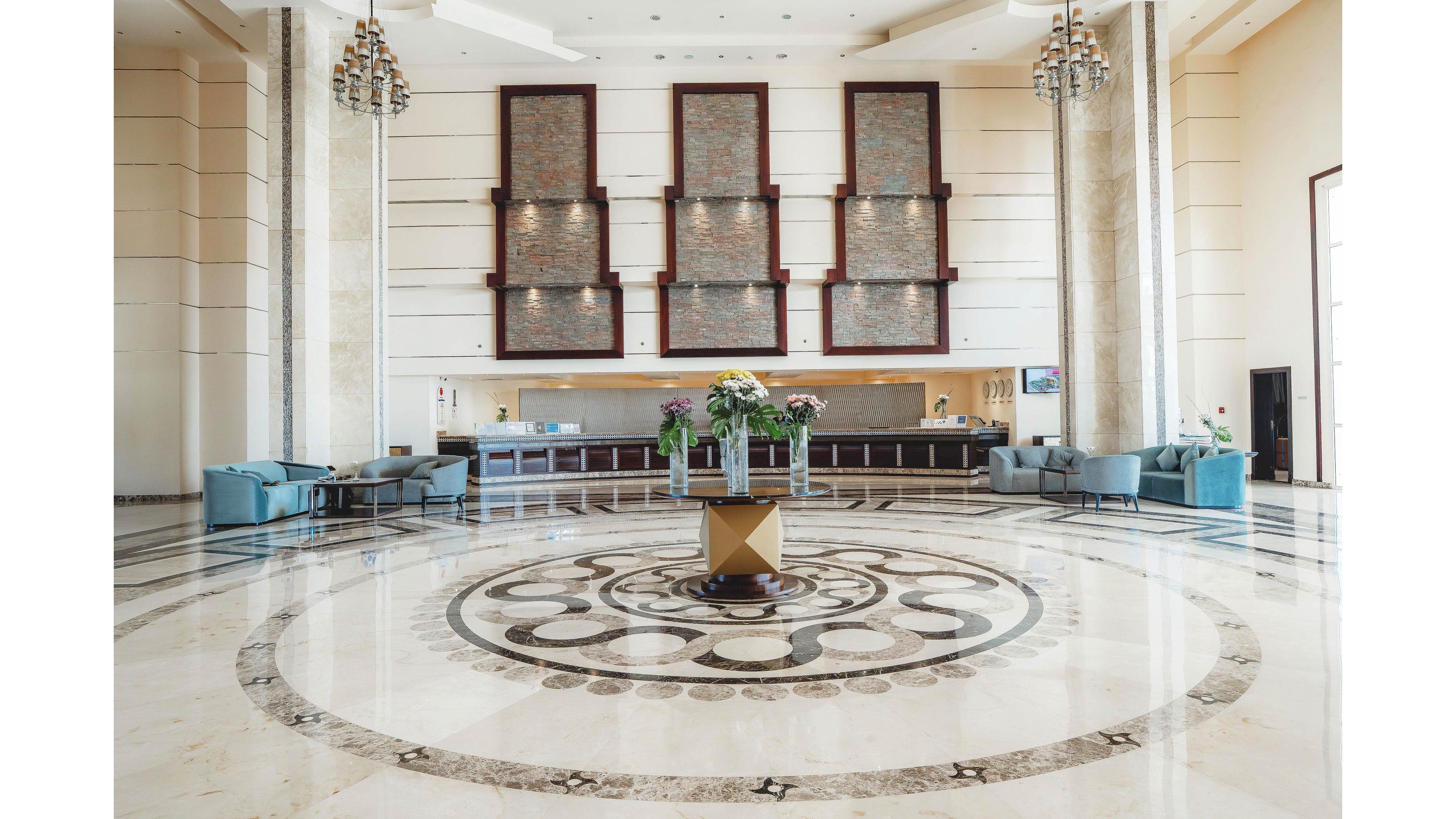 A large, elegant hotel lobby features a polished marble floor with an ornate circular pattern in the center. A floral arrangement on a pedestal stands as the focal point of the space. The reception desk stretches across the back wall beneath three tall decorative panels. Soft seating areas with armchairs and side tables are arranged along the sides, creating a welcoming and luxurious atmosphere.