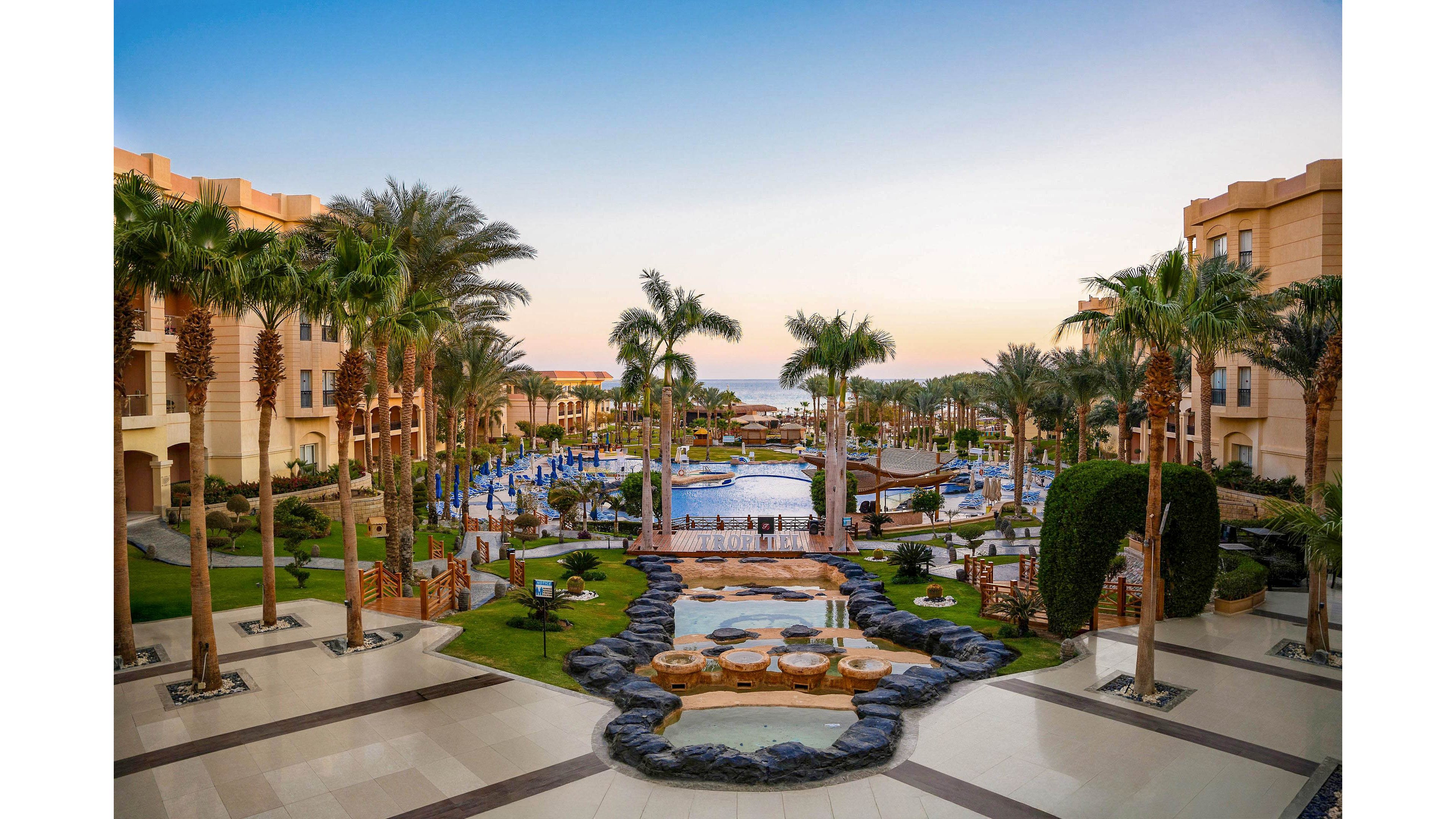 A large resort courtyard features multiple swimming pools surrounded by palm trees and sun loungers. Beige hotel buildings frame the scene on both sides, leading the eye toward the distant sea on the horizon. Pathways, wooden railings, and manicured gardens create a structured, inviting layout. The warm evening light and clear sky give the setting a calm, relaxing vacation atmosphere.