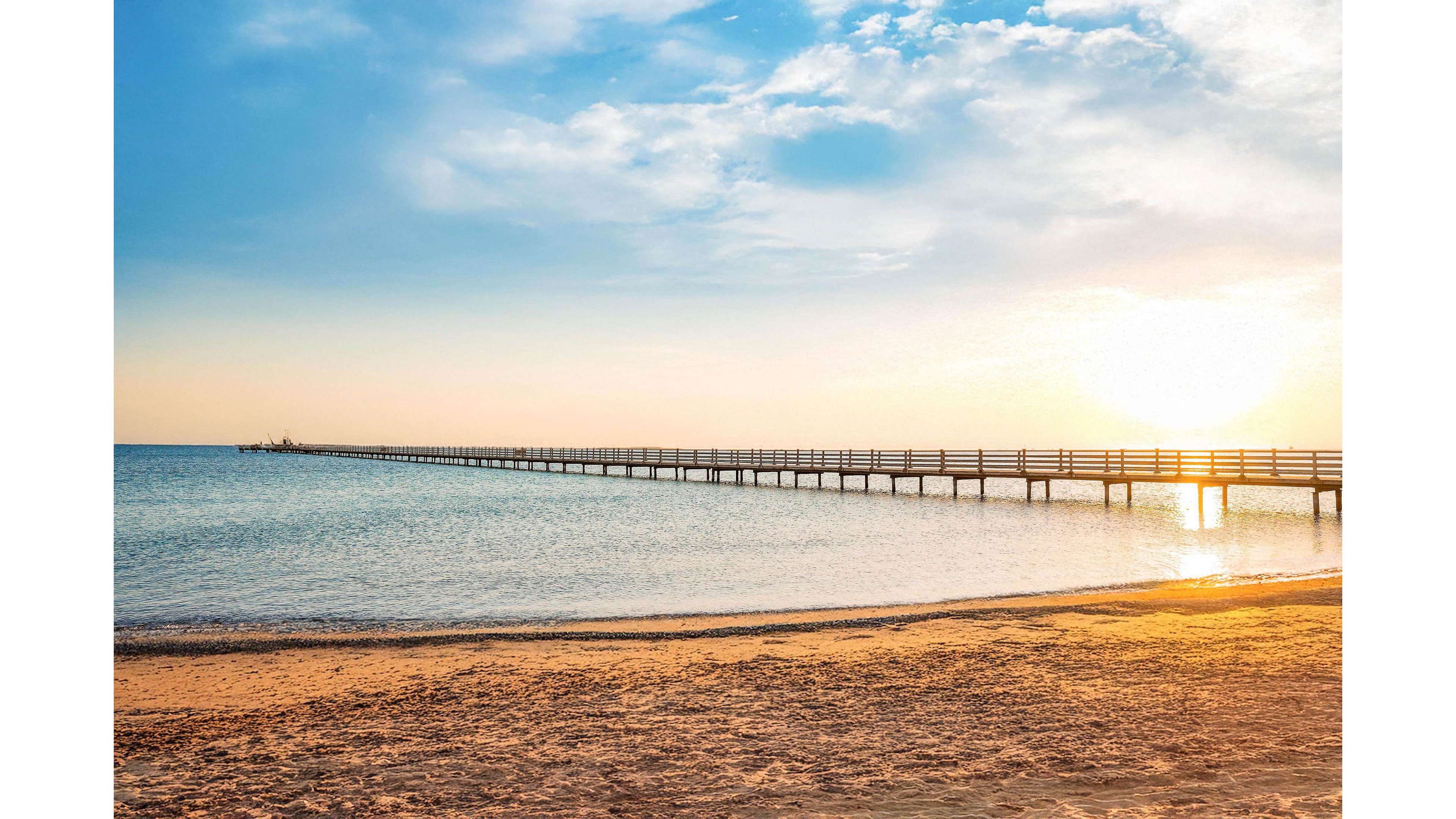 A calm seascape shows a long wooden pier stretching far into the water under a bright rising sun. Gentle waves lap against the sandy beach in the foreground. The sky is partly cloudy with soft blue and golden tones reflecting on the sea. The overall scene feels peaceful and serene, evoking a quiet early morning by the coast.
