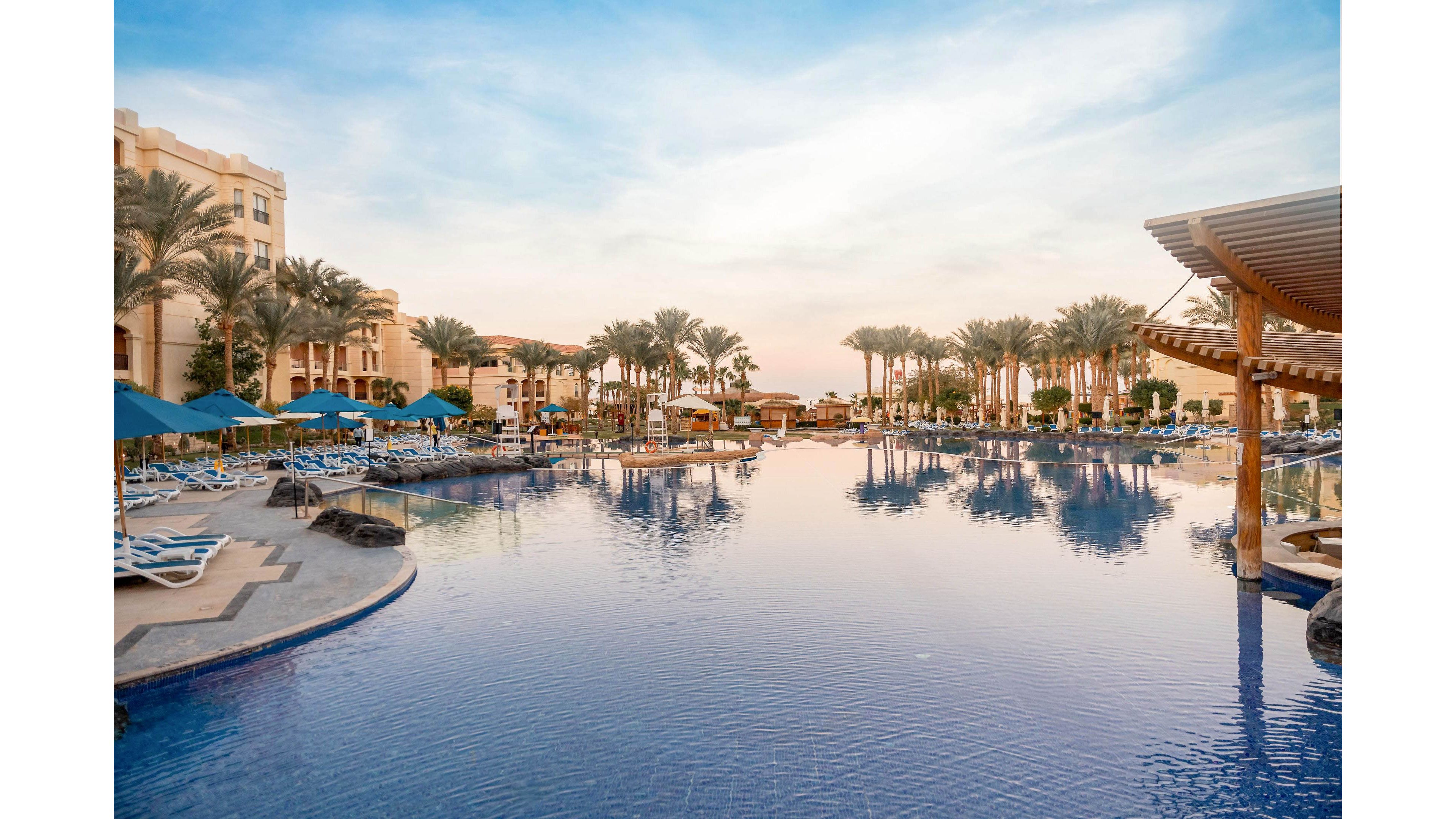 A large resort swimming pool stretches across the foreground, reflecting the soft colors of the evening sky. Sun loungers and blue umbrellas line the poolside, with a few guests relaxing near the water. Tall palm trees frame the scene and lead the eye toward the multi-story hotel buildings in the background. The calm water and warm light create a tranquil vacation atmosphere.