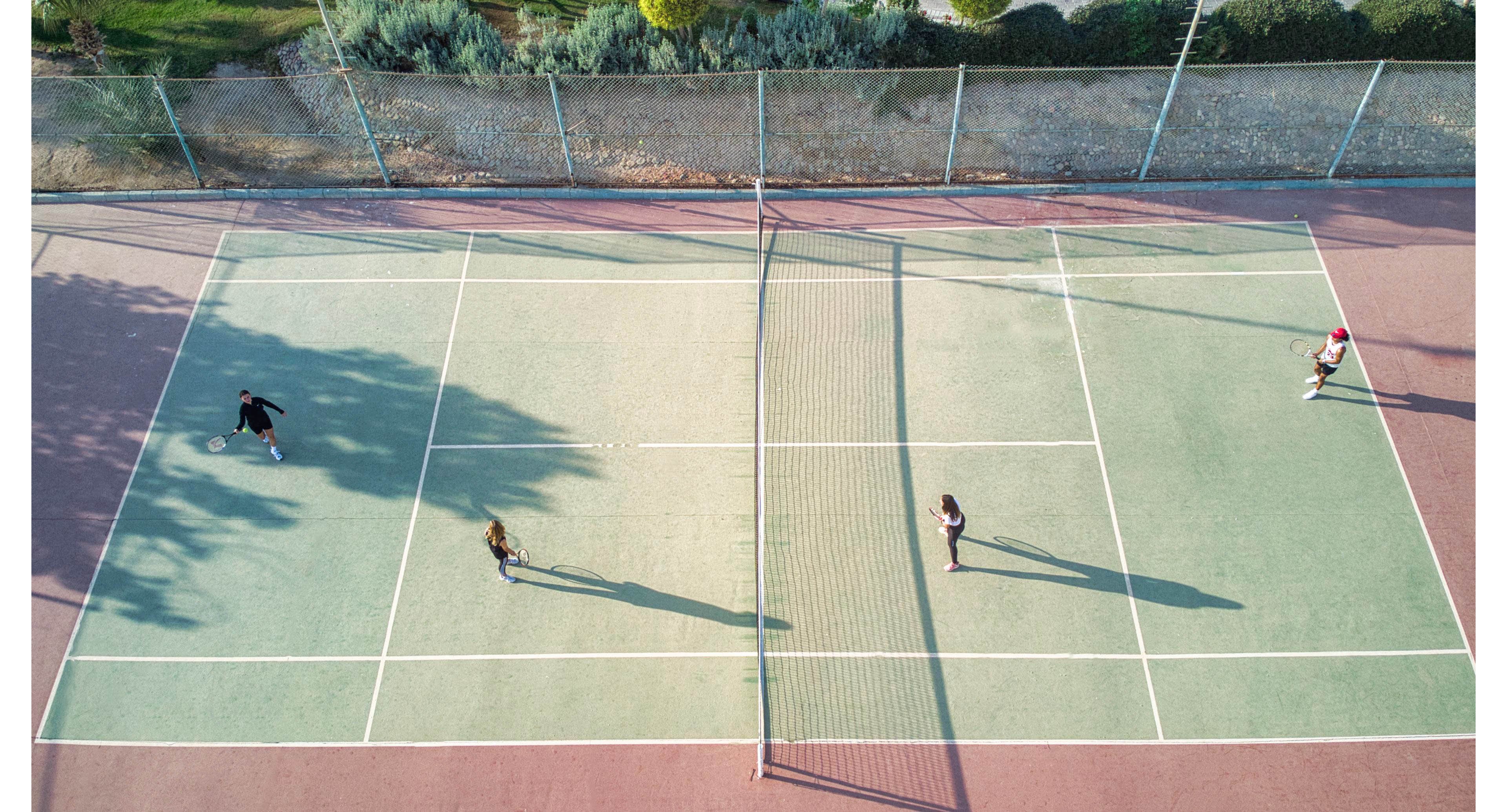 An overhead drone view shows four people on an outdoor tennis court. Two players stand on each side of the net, appearing to play a doubles match. Long shadows stretch across the court, suggesting late afternoon sunlight. The surrounding area includes fencing and greenery beyond the court surface.