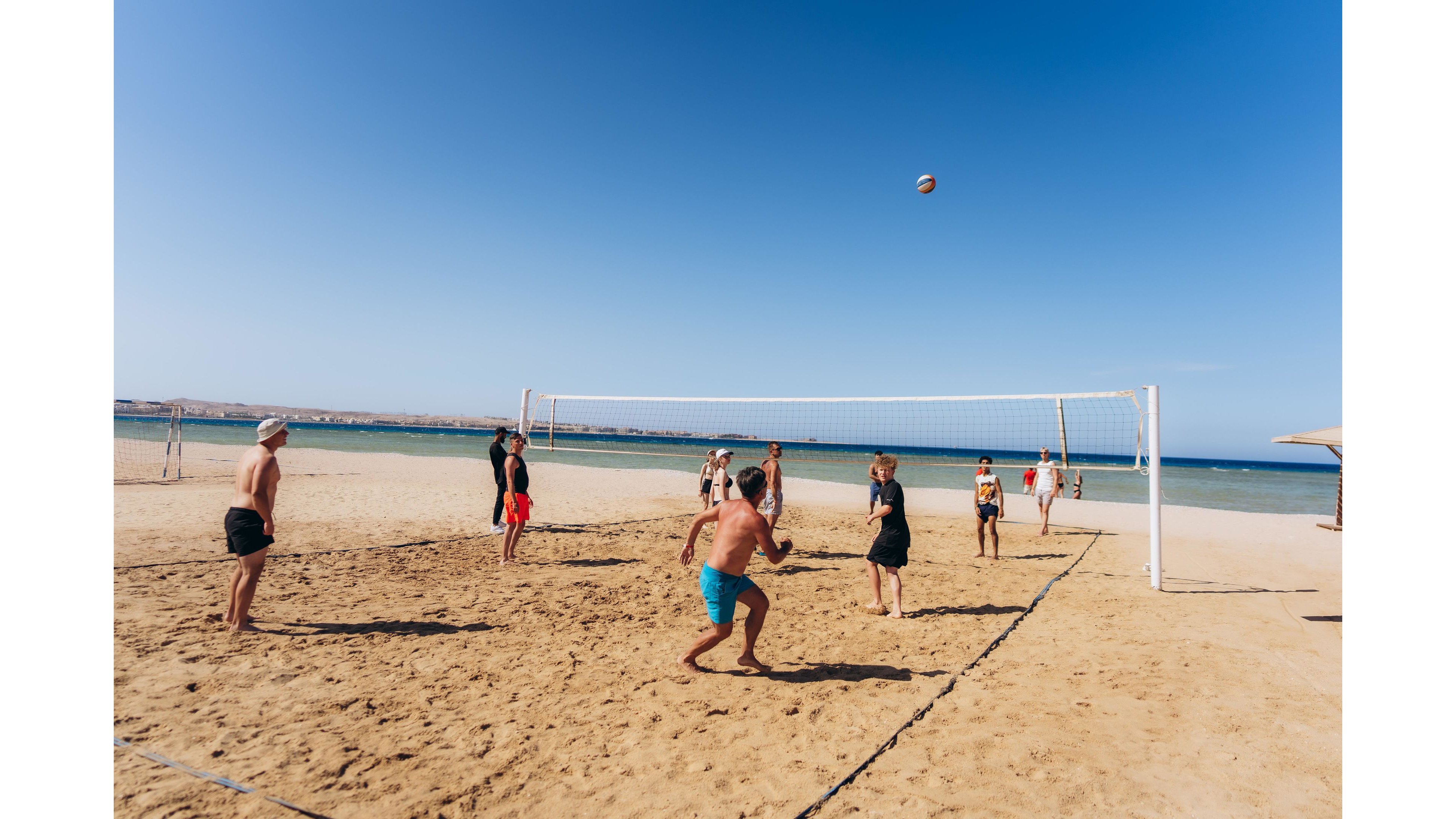 A group of people play volleyball on a sandy beach under a clear blue sky. The volleyball net is set up near the shoreline with the sea visible in the background. Players in swimwear are actively engaged in the game, jumping and preparing to hit the ball. The bright sunlight and open seaside setting create a relaxed, summery atmosphere.