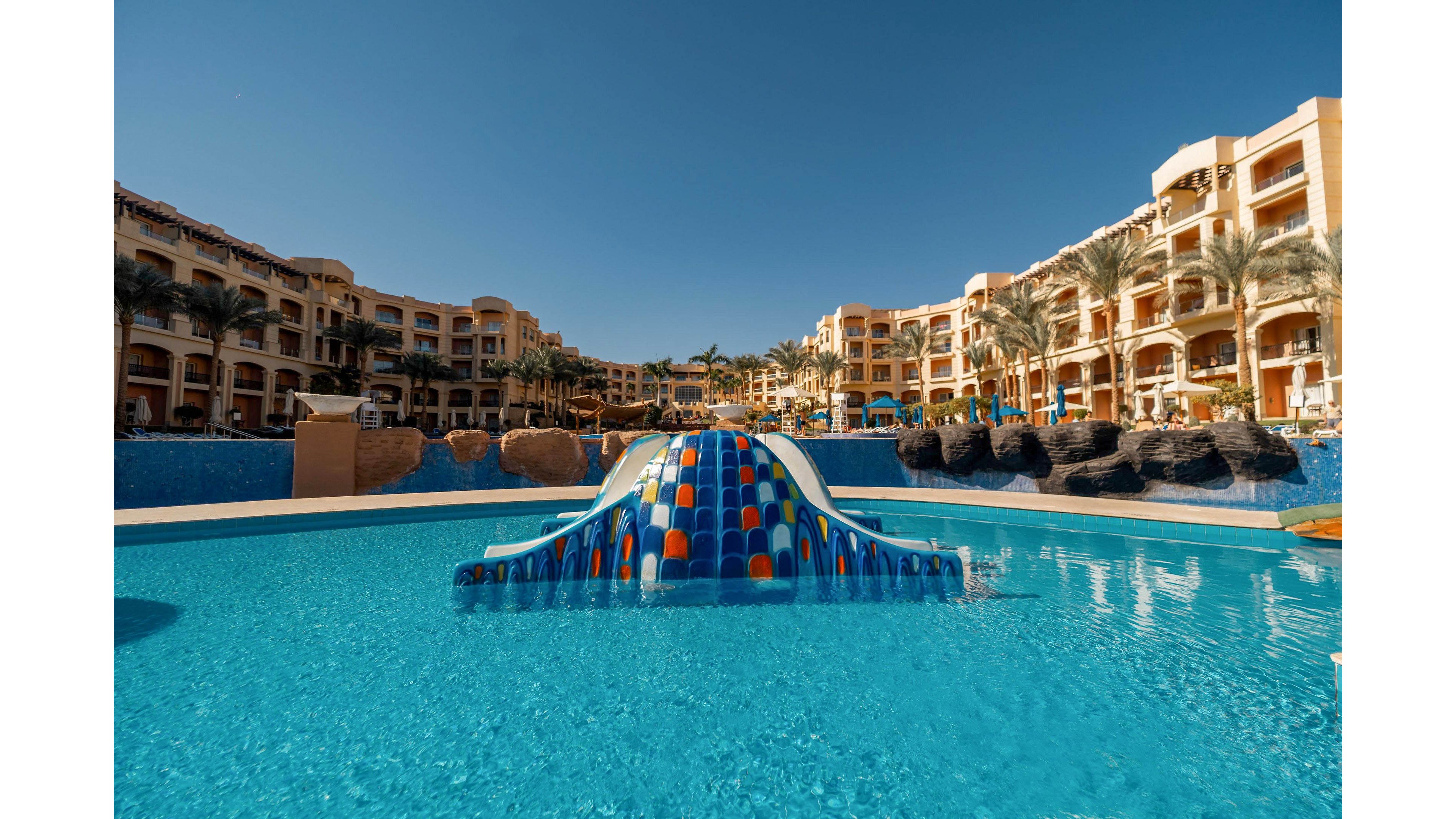 A large resort swimming pool features a colorful, curved water slide in the center. Beige multi-story hotel buildings with balconies surround the pool area under a clear blue sky. Palm trees and rock formations add a tropical atmosphere to the scene. The bright sunlight enhances the vivid blue water and warm tones of the architecture.