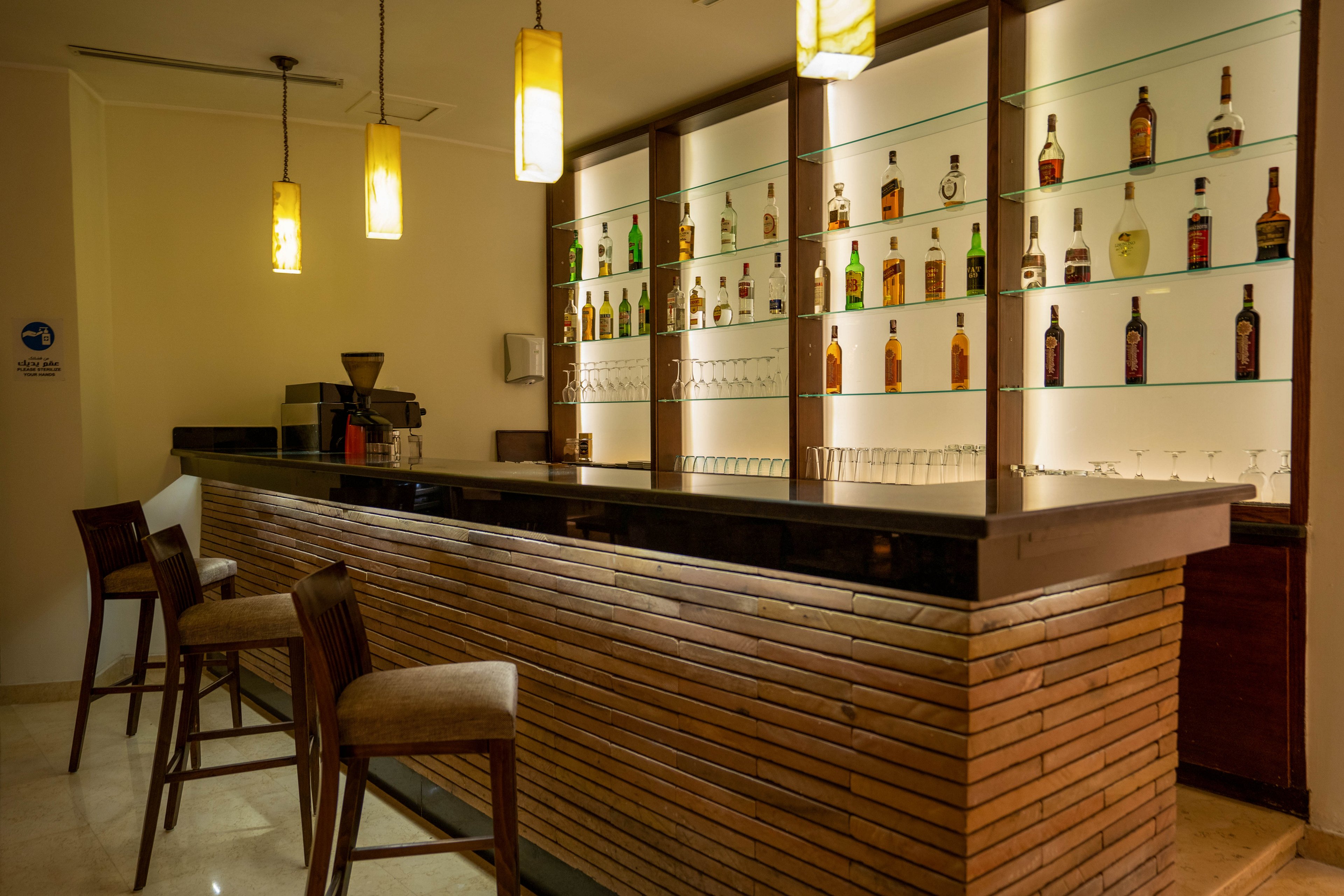 A contemporary bar features a sleek wooden counter and backlit shelves displaying various bottles of liquor. The setting is warmly lit with pendant lights, creating a cozy and inviting atmosphere. Three upholstered bar stools are arranged along the counter, and glassware is neatly organized on the shelves. The overall mood is relaxed and sophisticated, ideal for social gatherings or quiet evenings.
