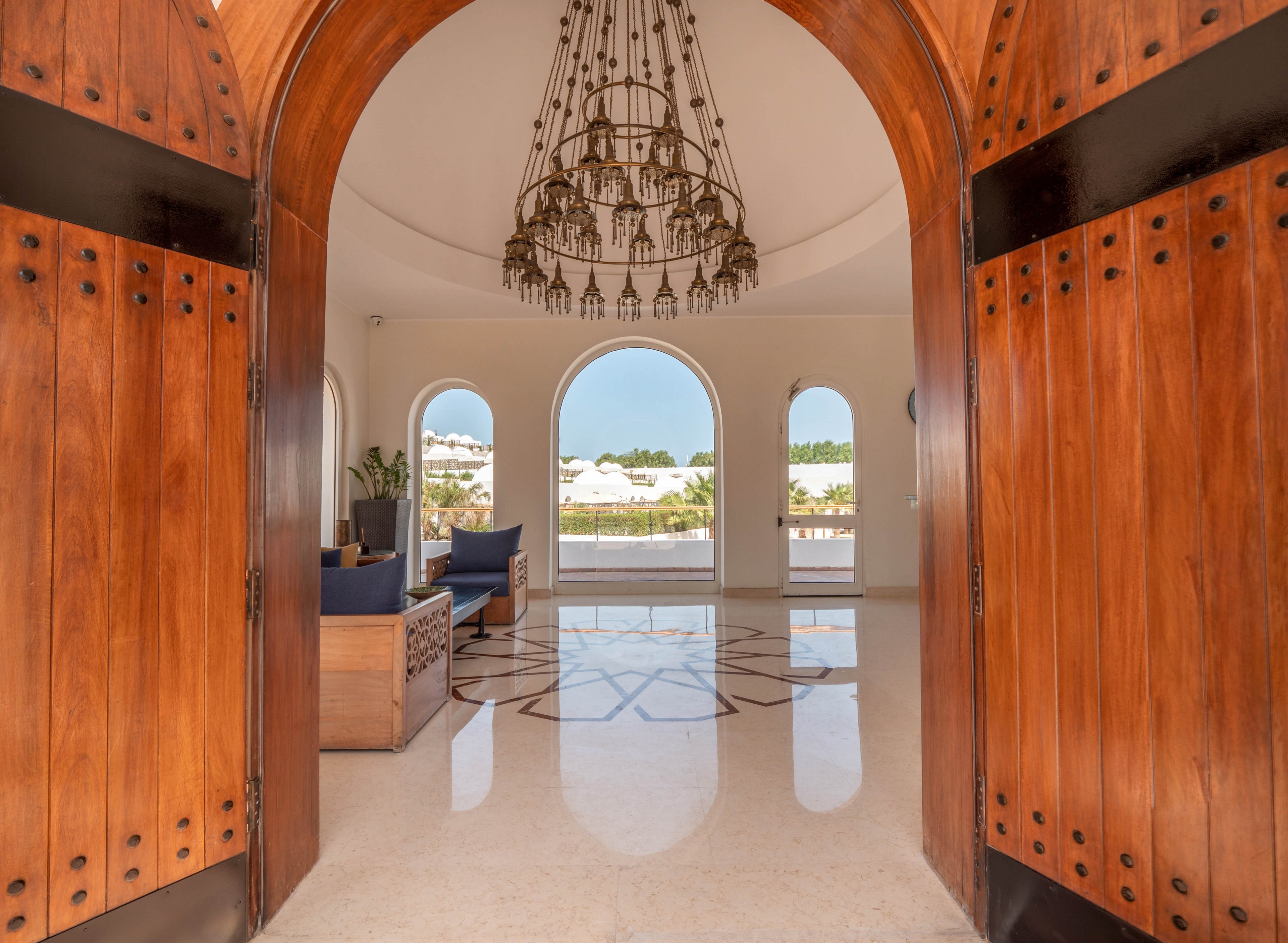Large arched wooden doors open to a bright, spacious room with polished floors and a grand chandelier. The interior features modern seating and large arched windows offering views of white buildings and greenery outside. The setting is airy and inviting, with natural light flooding the space and geometric floor patterns adding visual interest.
