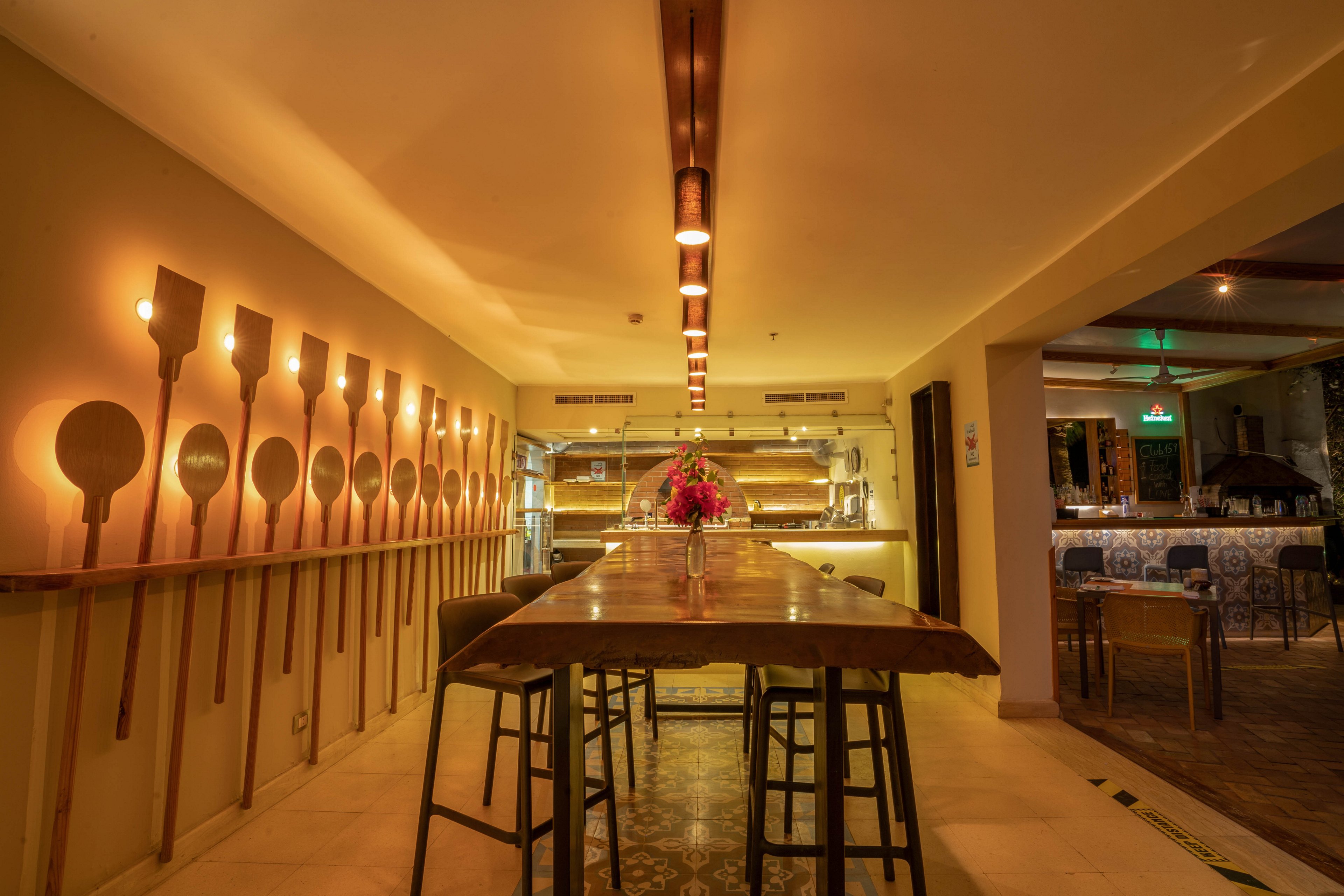 A contemporary restaurant interior features a long wooden communal table with stools, illuminated by a row of ceiling lights. The setting is cozy and inviting, with pizza peels mounted decoratively on the wall and a flower arrangement as a centerpiece. The open kitchen is visible in the background, and the overall palette is warm with soft yellow and brown tones. The atmosphere suggests a relaxed dining experience in a stylish, modern space.