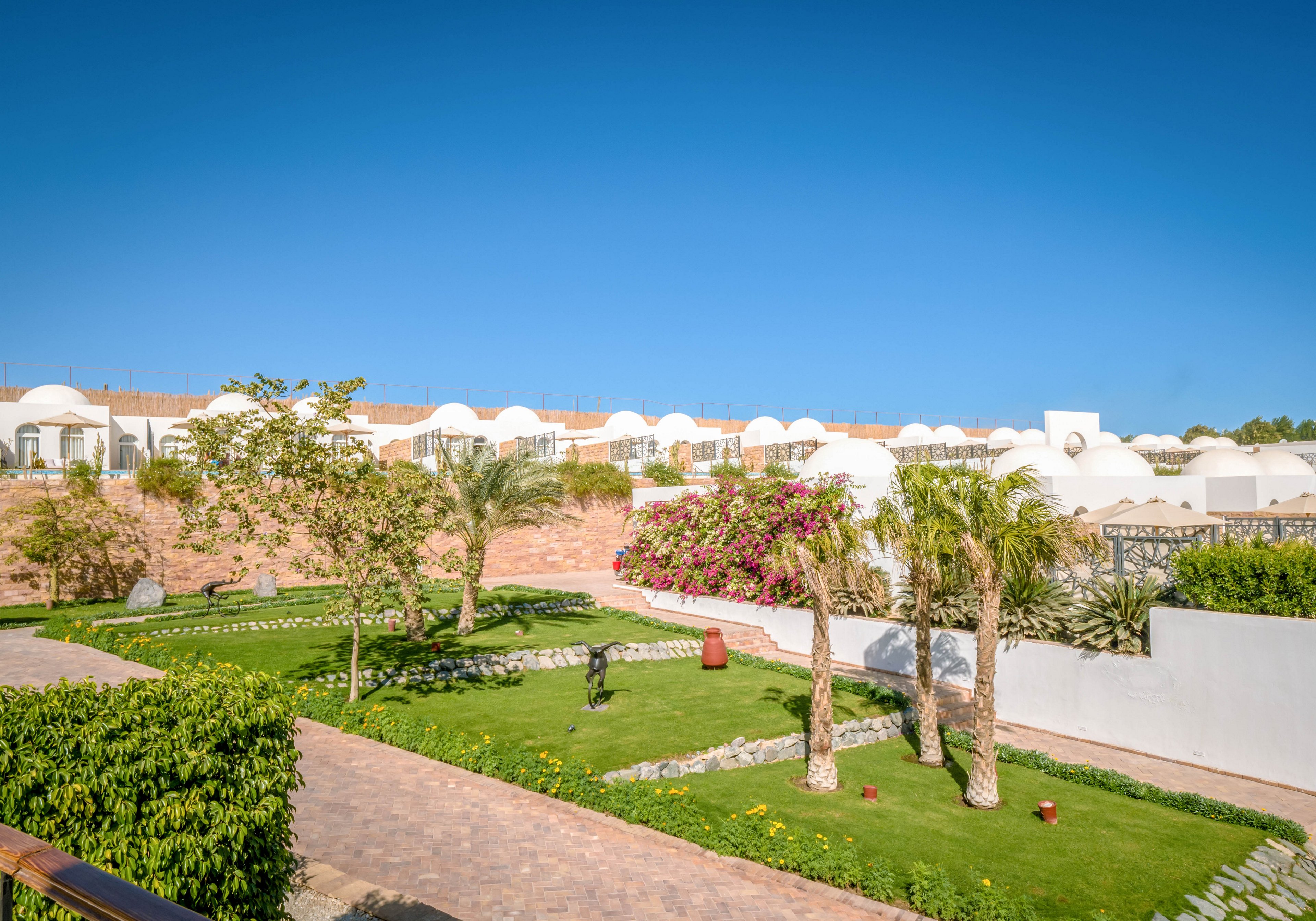 A lush green garden with palm trees and flowering bushes is set against a backdrop of white dome-shaped villas. The scene is bathed in bright sunlight under a clear blue sky, creating a tranquil and inviting atmosphere. Pathways wind through the landscaped grounds, highlighting the resort's relaxing outdoor setting. No visible text or numbers are present in the image.