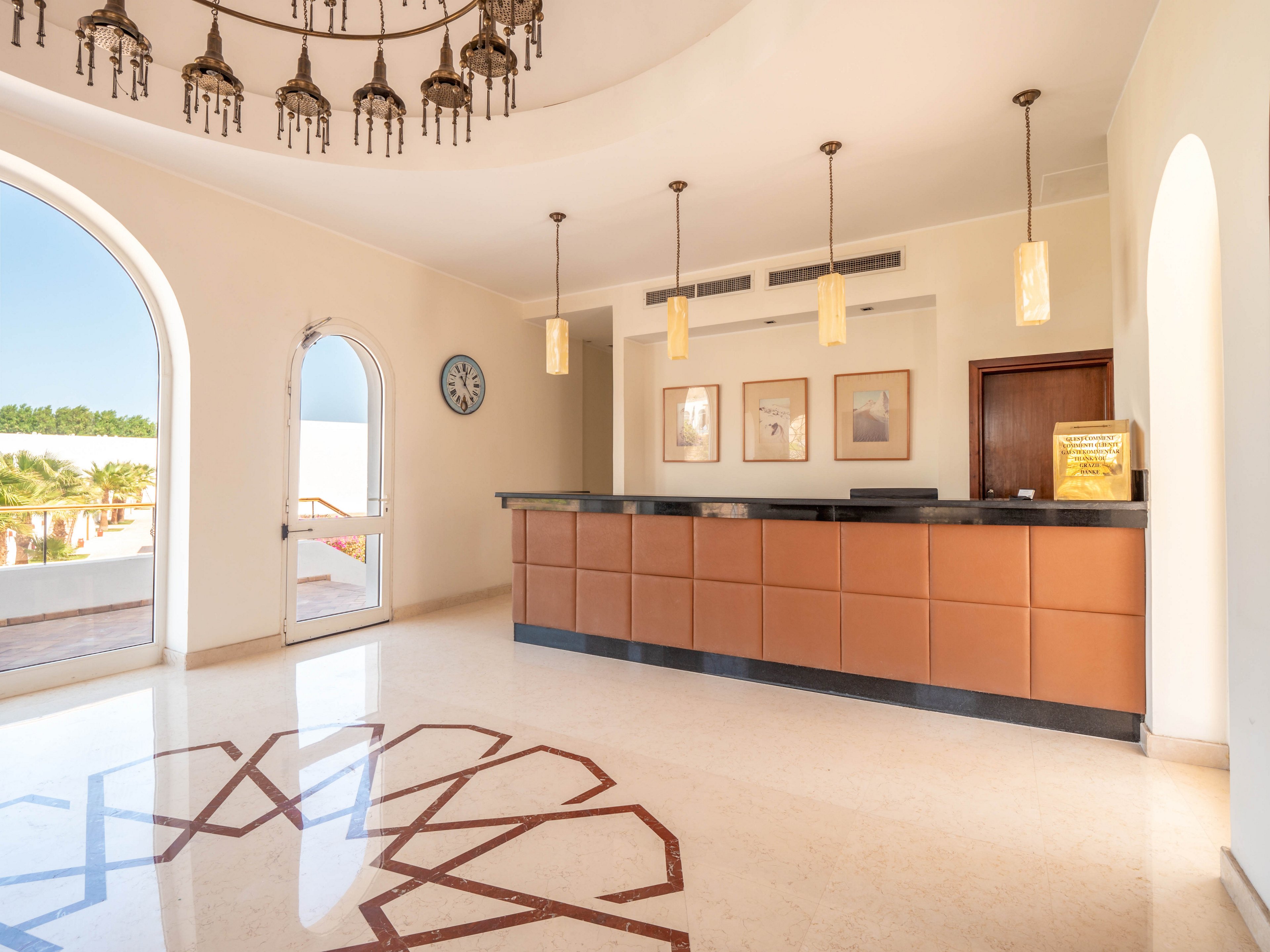 A spacious hotel reception area features a large brown front desk and modern pendant lighting. The setting is bright with natural light streaming through arched windows, highlighting a geometric floor pattern. Wall art and a visible clock add to the welcoming atmosphere. The overall palette is neutral with warm accents, creating a calm and inviting mood.