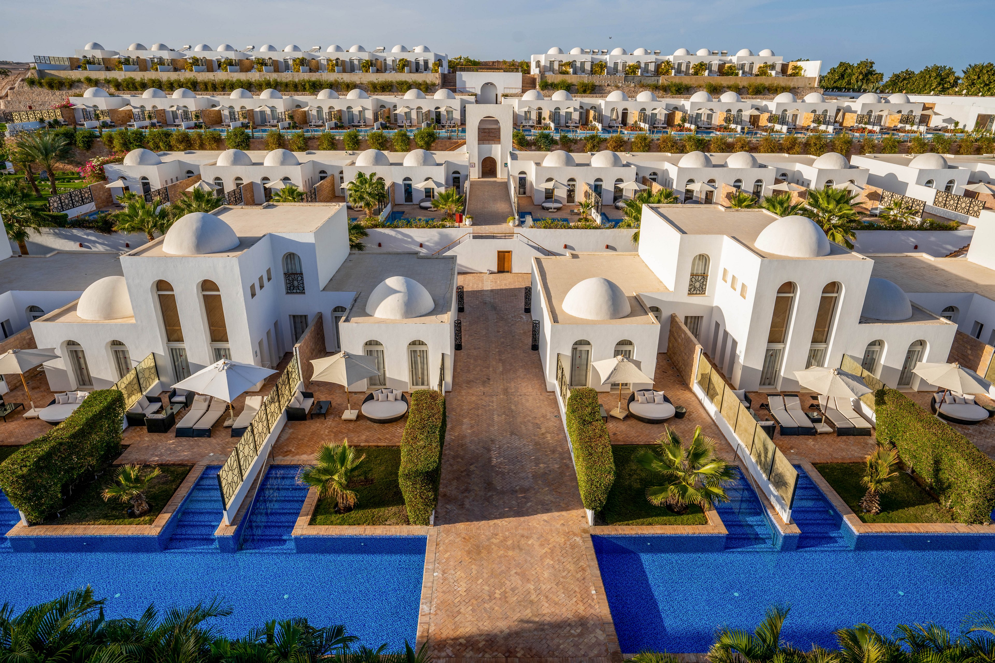 A series of white, dome-roofed villas are arranged in symmetrical rows, each with private terraces and direct access to a blue swimming pool. The architecture features clean lines and arches, evoking a Mediterranean or Middle Eastern style. Palm trees and manicured hedges add a lush, inviting atmosphere. The setting is sunlit, creating a bright and tranquil mood. No visible text or numbers are present in the image.