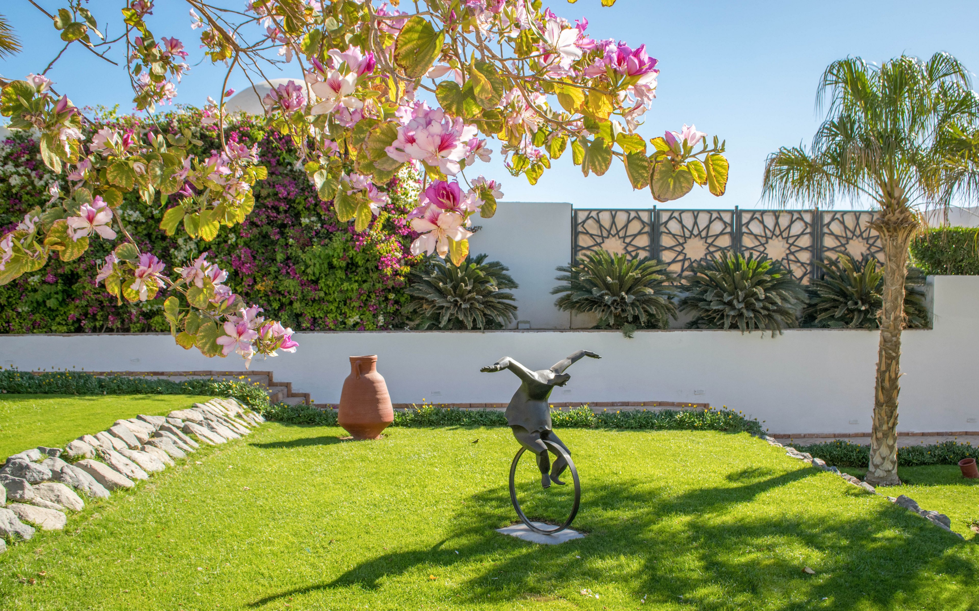 A lush green garden features a whimsical metal sculpture of a person balancing on a unicycle. The setting is bright and sunny, with a flowering tree in the foreground and a palm tree to the right. Decorative wall panels and a large terracotta pot add visual interest to the landscaped space. The overall mood is cheerful and tranquil, with vibrant colors and clear blue sky.