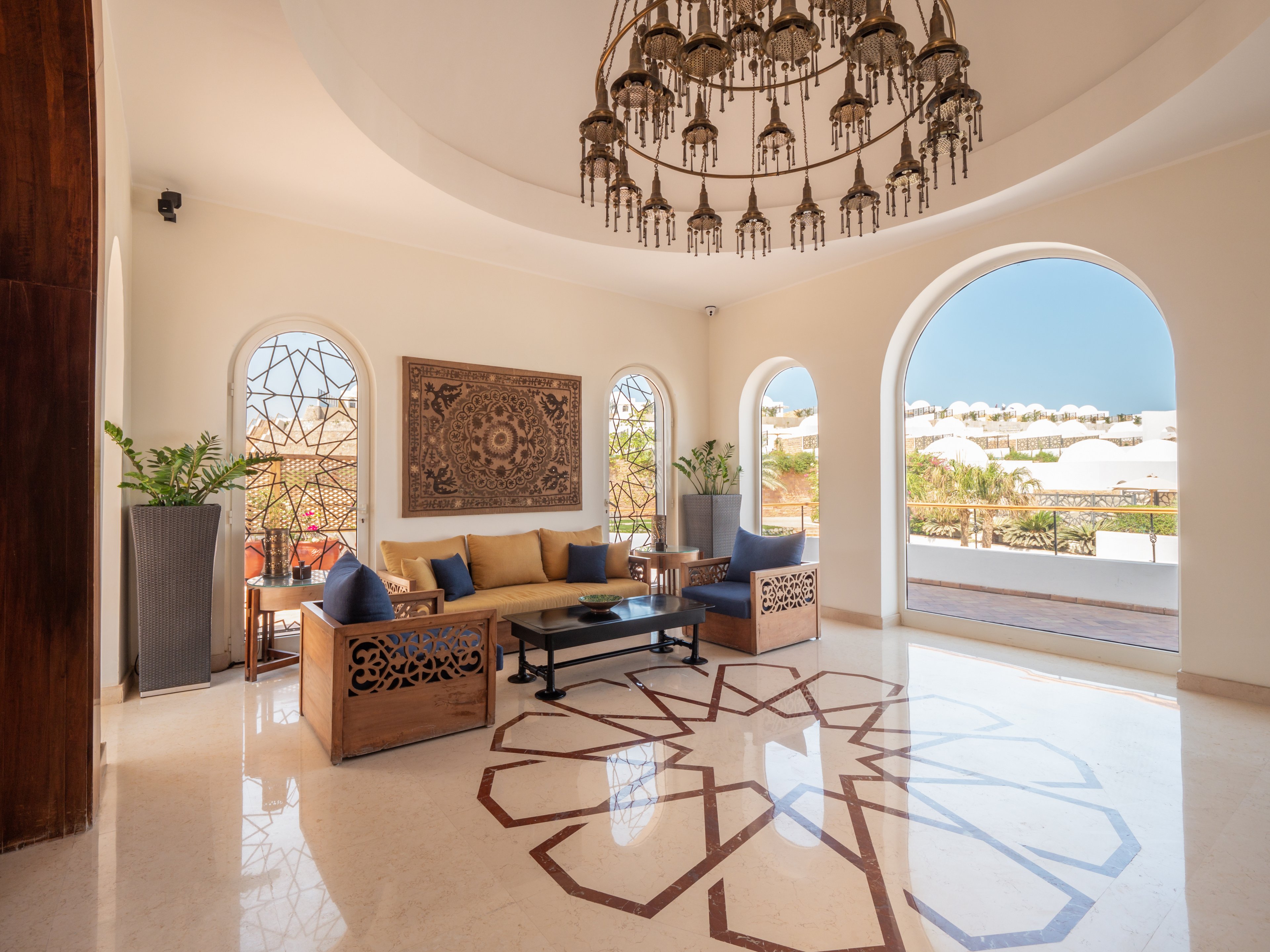 A spacious, sunlit lounge features a large decorative chandelier and intricate wall art. The room is furnished with wooden sofas and a central coffee table, set atop a polished floor with a geometric inlay. Large arched windows offer views of a bright outdoor terrace and distant white buildings. The setting is airy and inviting, with neutral tones and subtle blue accents. Potted plants add a touch of greenery to the sophisticated, Mediterranean-inspired space.