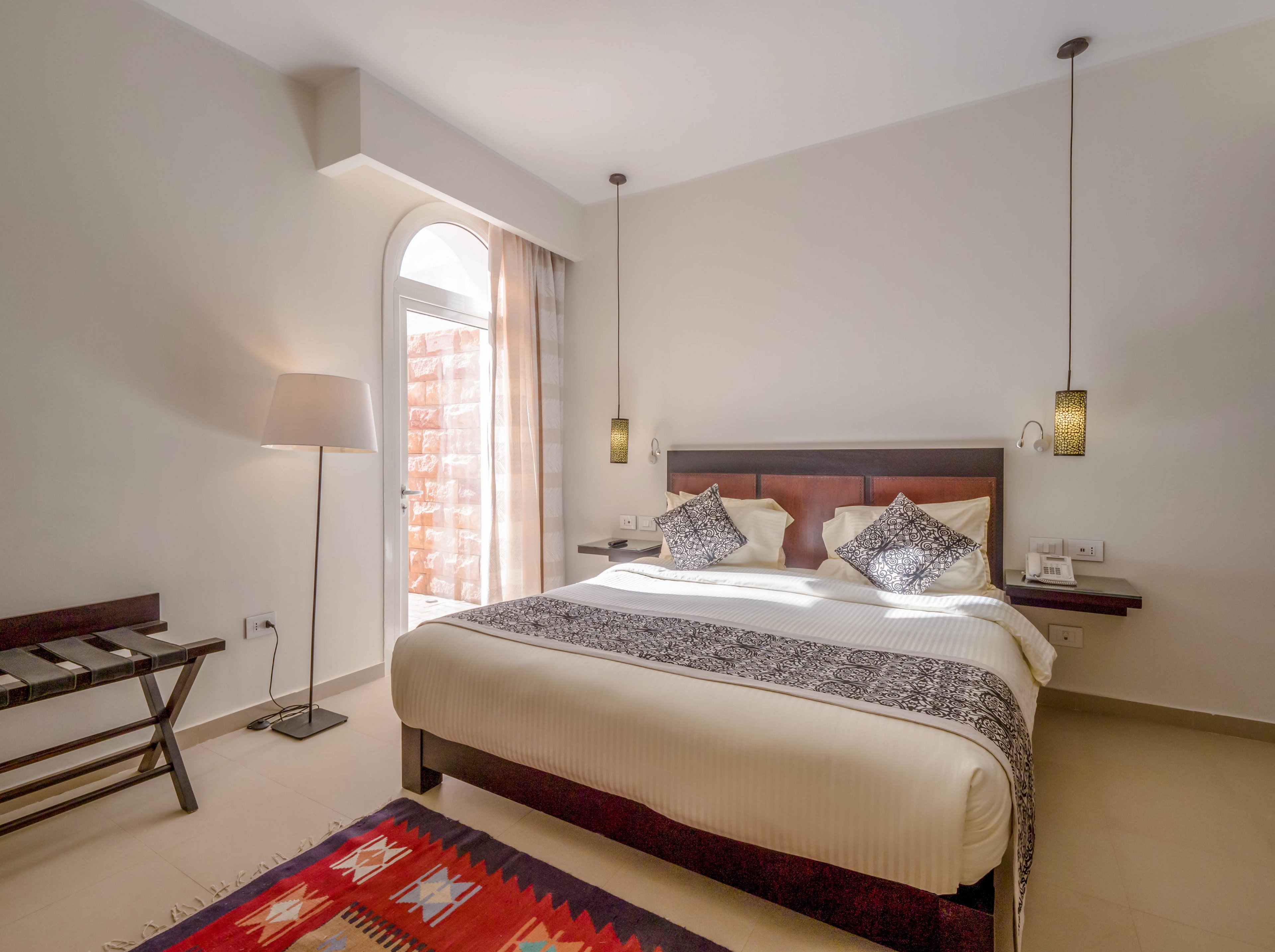 A neatly arranged hotel bedroom features a large double bed with patterned pillows and a decorative runner. The room is illuminated by natural light from a tall arched window, complemented by pendant lights and a standing lamp. Minimalist furniture, including a luggage rack and floating nightstands, enhances the clean, modern aesthetic. A colorful geometric rug adds warmth to the neutral palette. No visible text or numbers are present in the image.