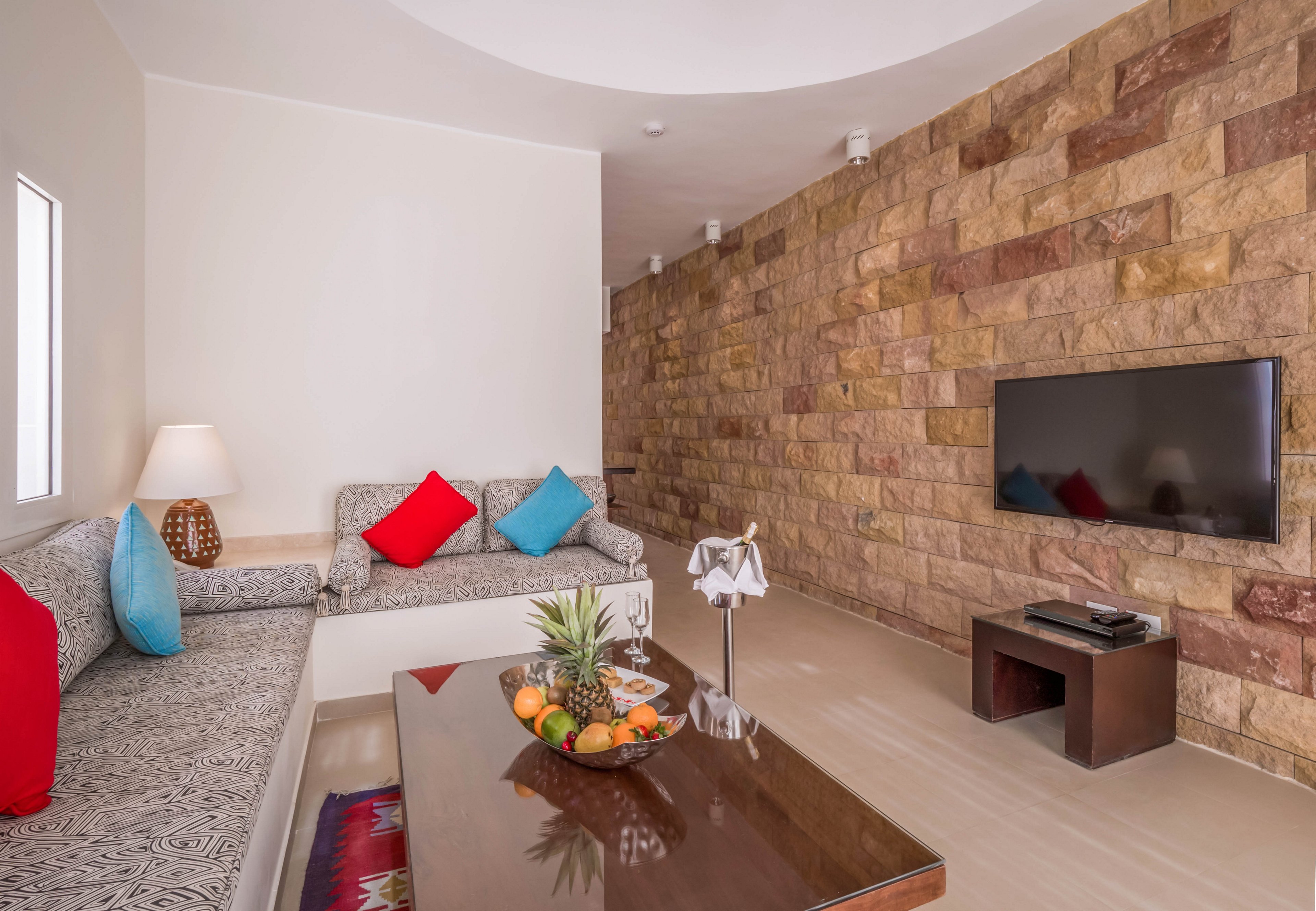 A contemporary living room features a patterned sectional sofa with colorful red and blue accent pillows. The space includes a sleek wooden coffee table topped with a fruit arrangement and a decorative napkin holder. A flat-screen television is mounted on a textured stone accent wall, with a media console below. The room is well-lit with natural light from a window and a table lamp, creating a cozy and inviting atmosphere.