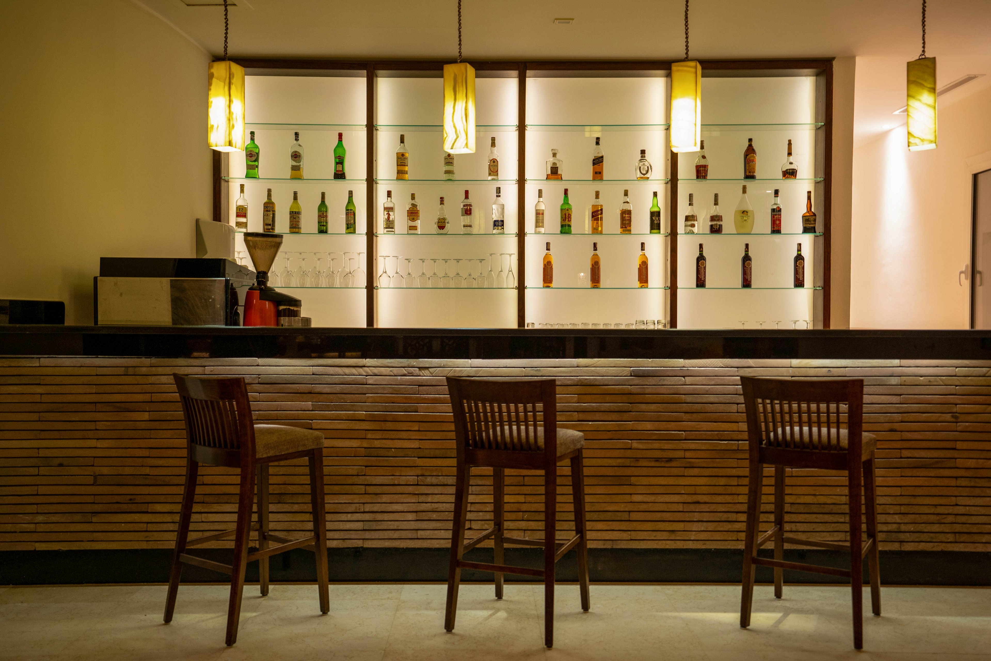 A contemporary bar interior features a wooden counter and three empty bar stools. The back wall displays a variety of bottles on illuminated shelves, creating a warm and inviting atmosphere. Three pendant lights hang above the bar, casting a soft yellow glow. The setting is clean and minimalist, with no people present. No visible text or numeric values are present in the image.