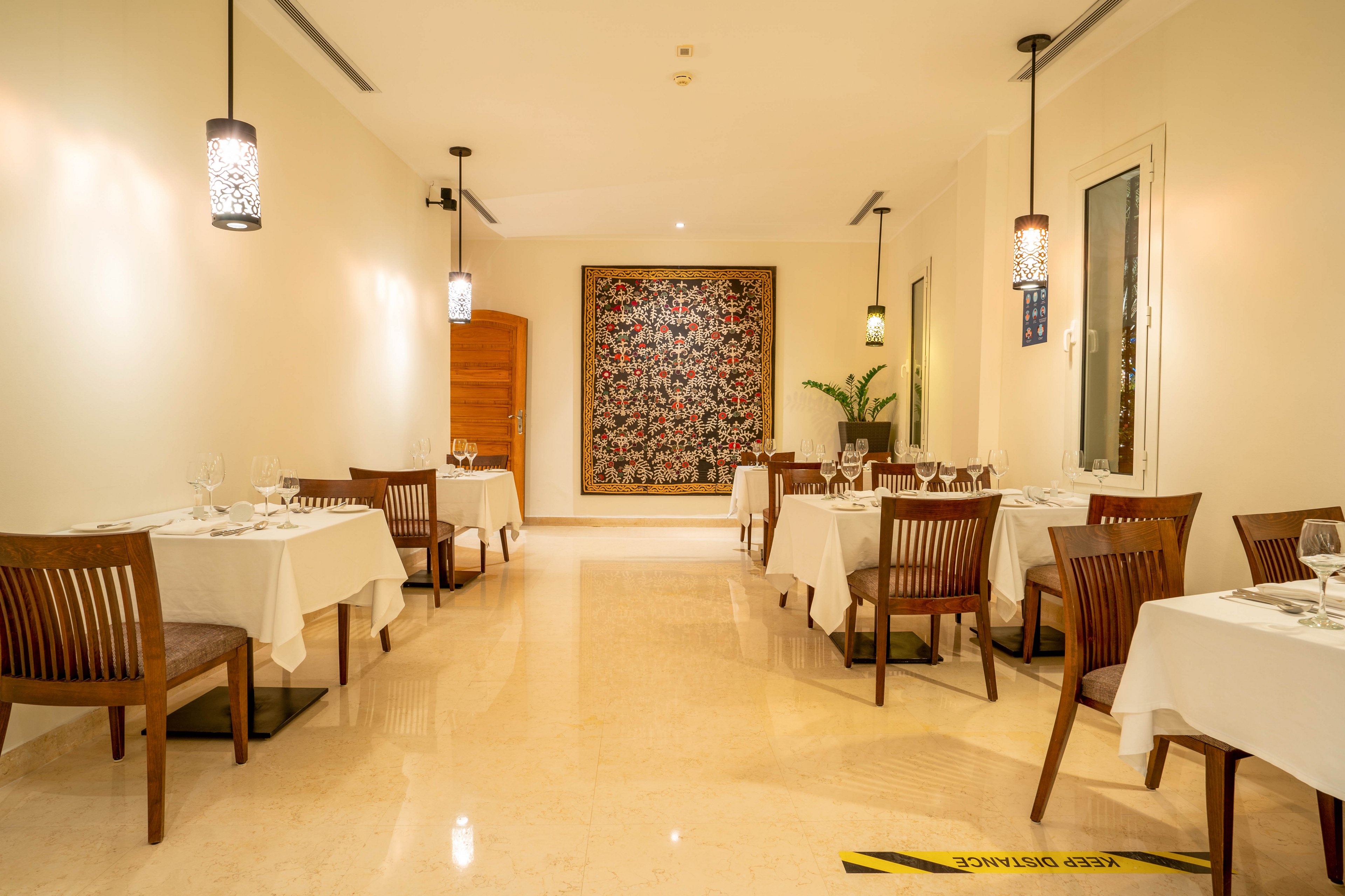A well-lit restaurant dining area features neatly arranged tables with white tablecloths and wooden chairs. The setting is modern and minimalistic, with pendant lights and a large decorative artwork on the back wall. The polished floor and neutral color palette create a calm, inviting atmosphere. A yellow caution sign with the text 'Caution Wet Floor' is visible on the floor.