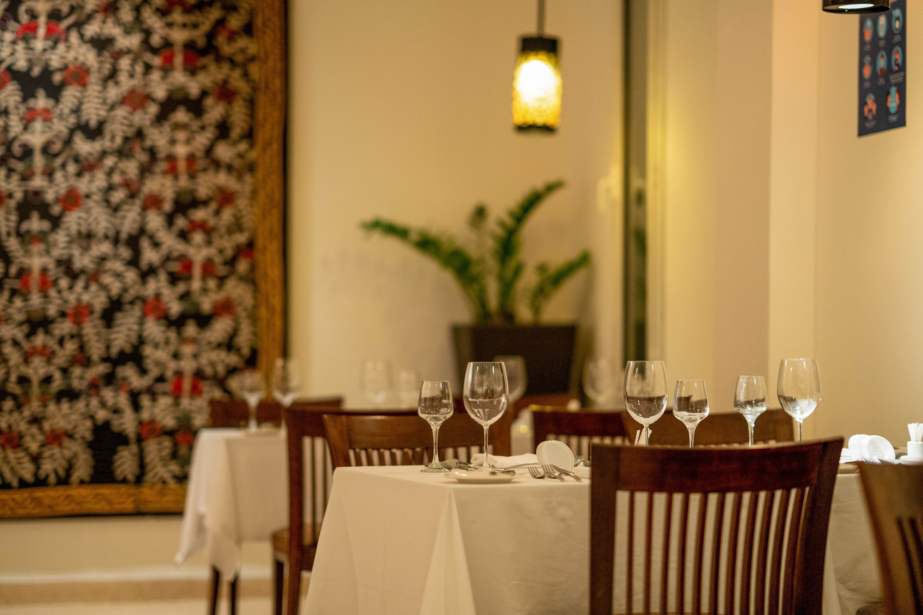 A cozy restaurant interior features neatly arranged tables covered with white tablecloths and set with multiple wine glasses and cutlery. The setting is softly lit with a warm hanging lamp and decorated with a patterned wall tapestry and a potted plant. The ambiance suggests a refined, intimate dining experience. No visible text or numbers are present in the image.