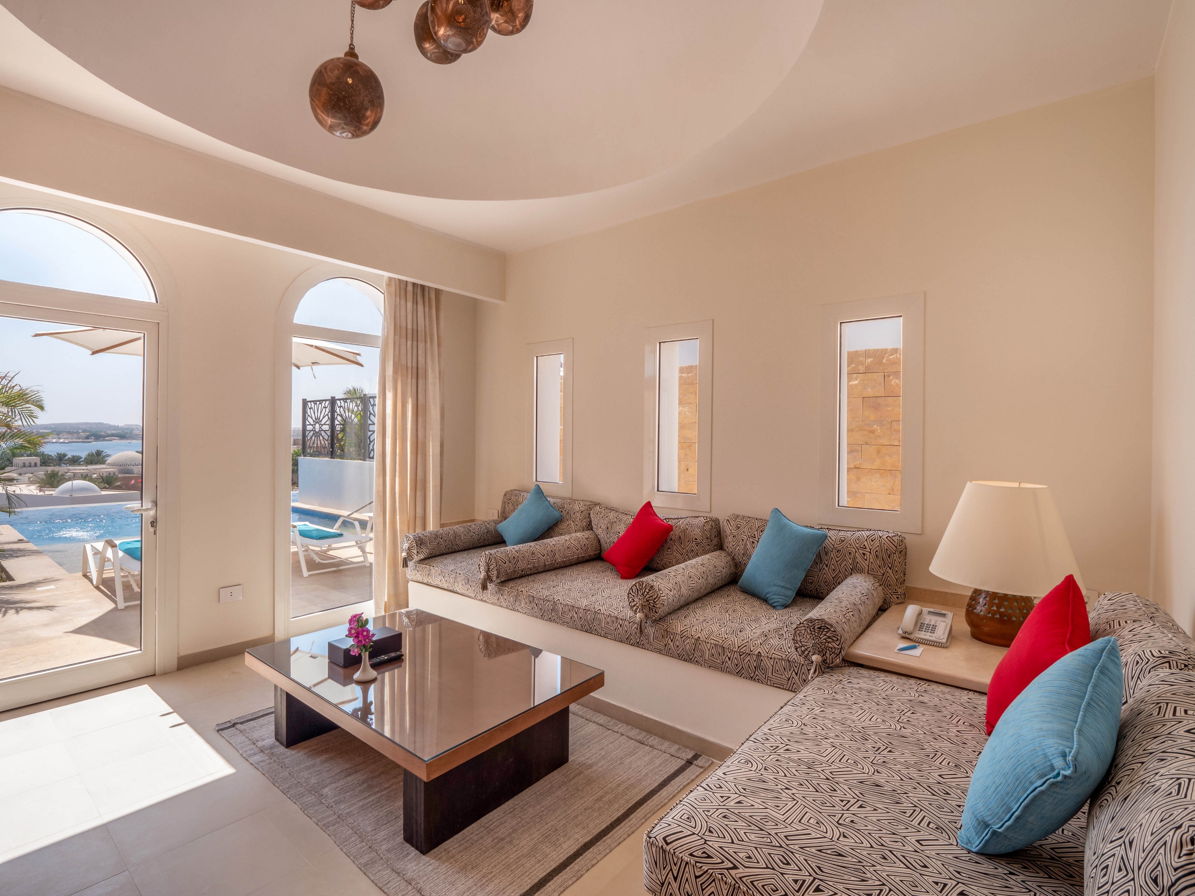 A sunlit living room features patterned sofas with colorful red and blue accent pillows, a modern coffee table, and a large lamp. Through arched glass doors, a private pool and outdoor lounge chairs are visible, suggesting a relaxing vacation setting. The neutral palette is accented by pops of color and natural light, creating a welcoming and airy atmosphere.