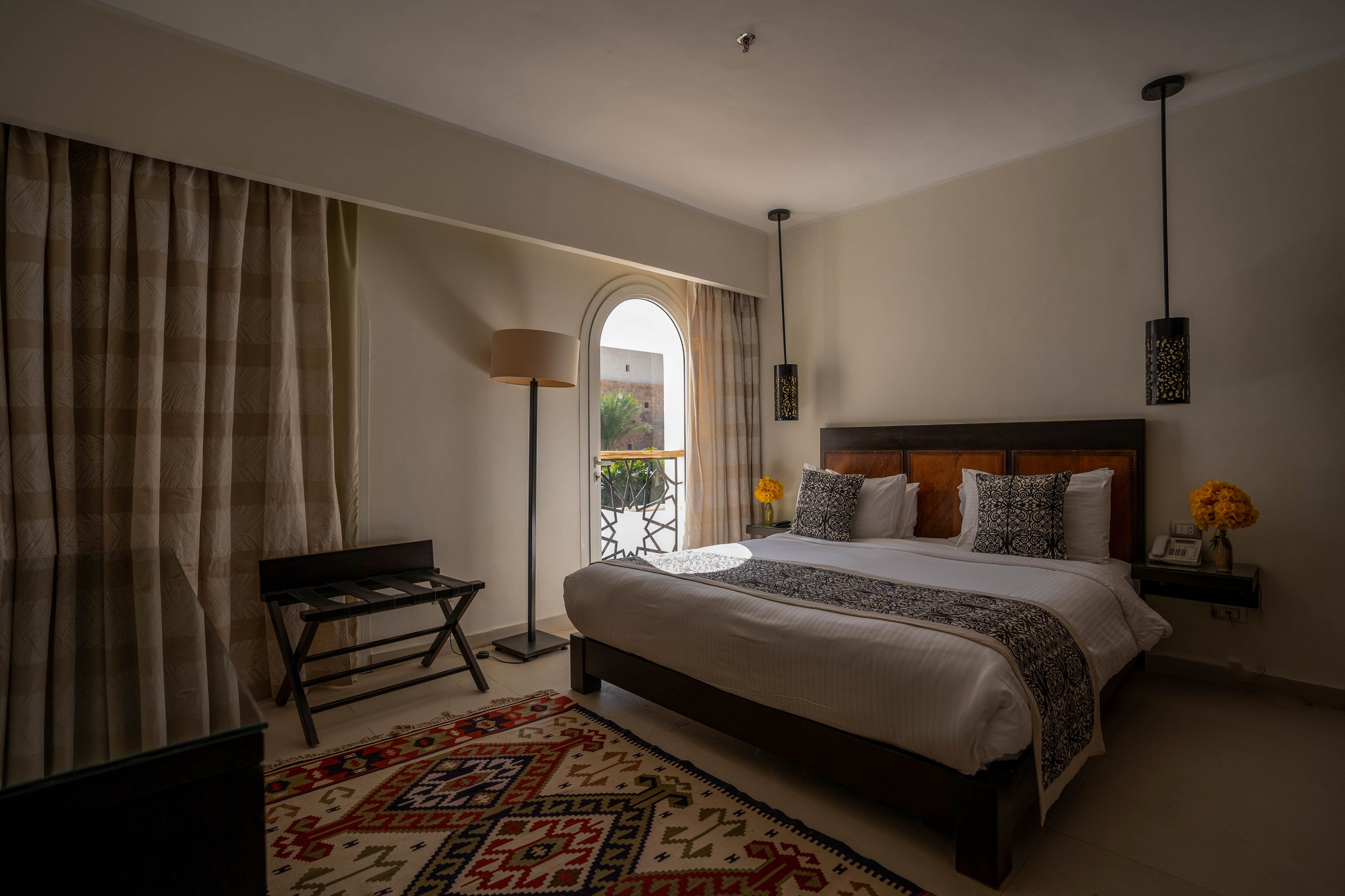 A contemporary hotel room features a large bed with decorative pillows and a patterned runner. The space is softly lit, with natural light streaming through an arched balcony door. Key visuals include a geometric rug, pendant lights, and yellow flowers on bedside tables, creating a cozy and inviting atmosphere. Neutral tones dominate the palette, with subtle accents adding warmth.