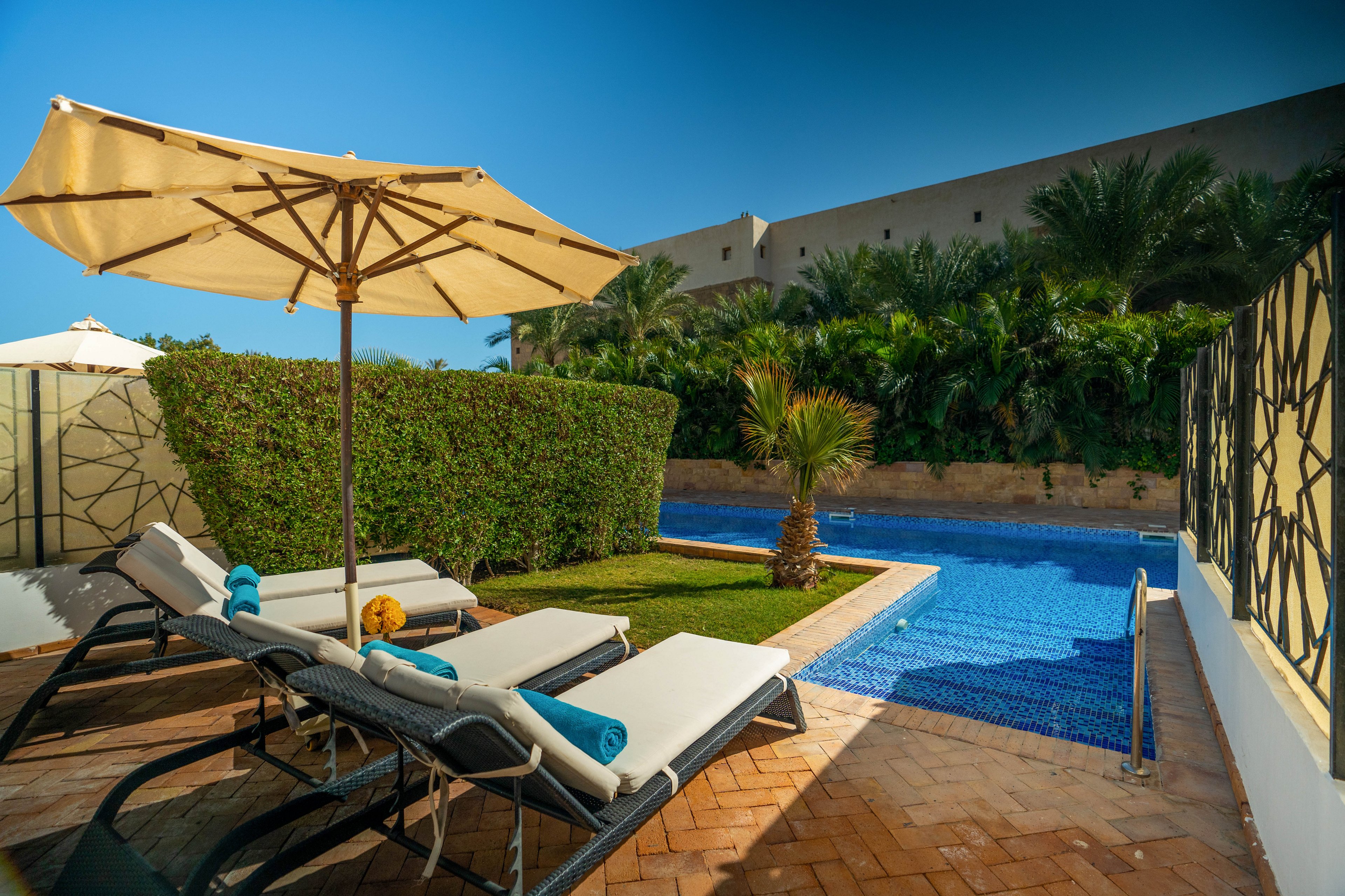 A tranquil outdoor pool area features two cushioned lounge chairs under a large beige umbrella. The setting is surrounded by lush green hedges and palm trees, with a clear blue swimming pool inviting relaxation. Rolled towels rest on the loungers, and the scene is bathed in bright sunlight, creating a serene and inviting atmosphere.