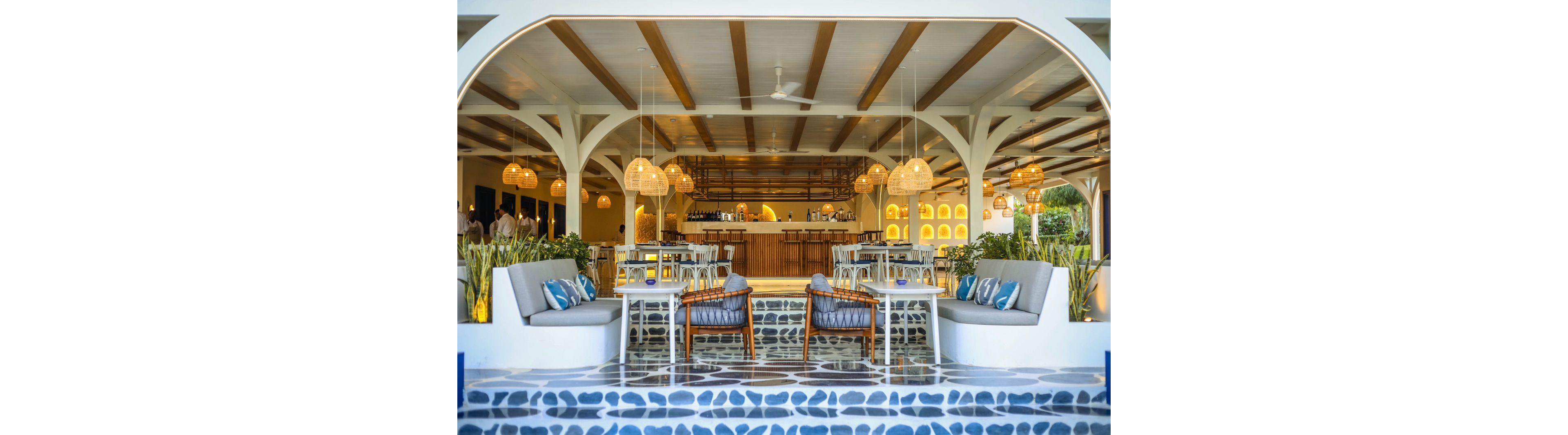 Stylish open-air bar lounge features cushioned chairs and sofas arranged around small tables. The setting appears tropical, with bright natural light and greenery framing the space. Warm pendant lights and wooden ceiling beams create a welcoming, contemporary coastal atmosphere. Blue and white patterned flooring and accent pillows add a fresh, nautical-inspired touch.