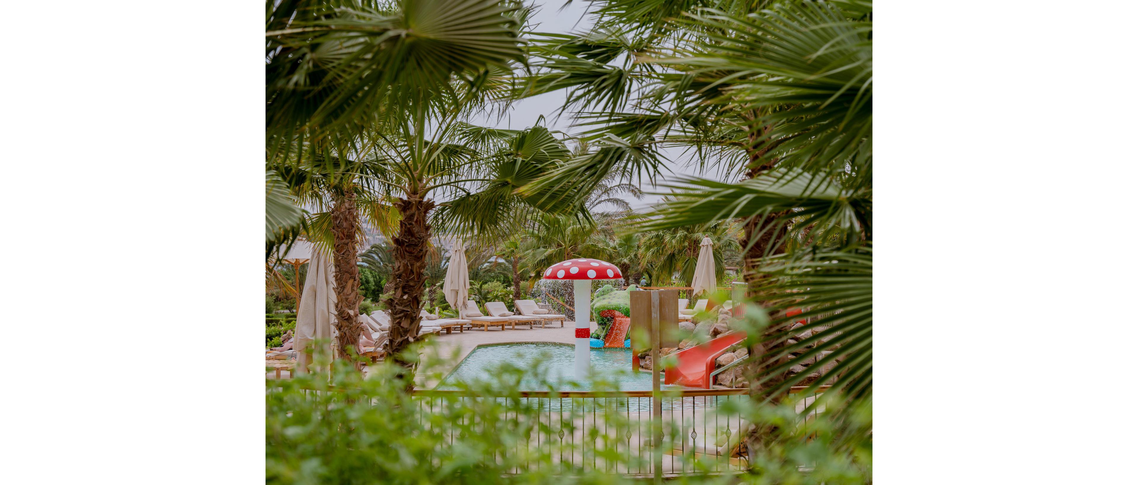 A colorful resort swimming pool is framed by lush palm trees and greenery. At the center stands a red and white mushroom-shaped water feature beside a bright red slide. Sun loungers and closed umbrellas line the poolside, suggesting a relaxing vacation setting. The overall scene feels tropical, family-friendly, and inviting.