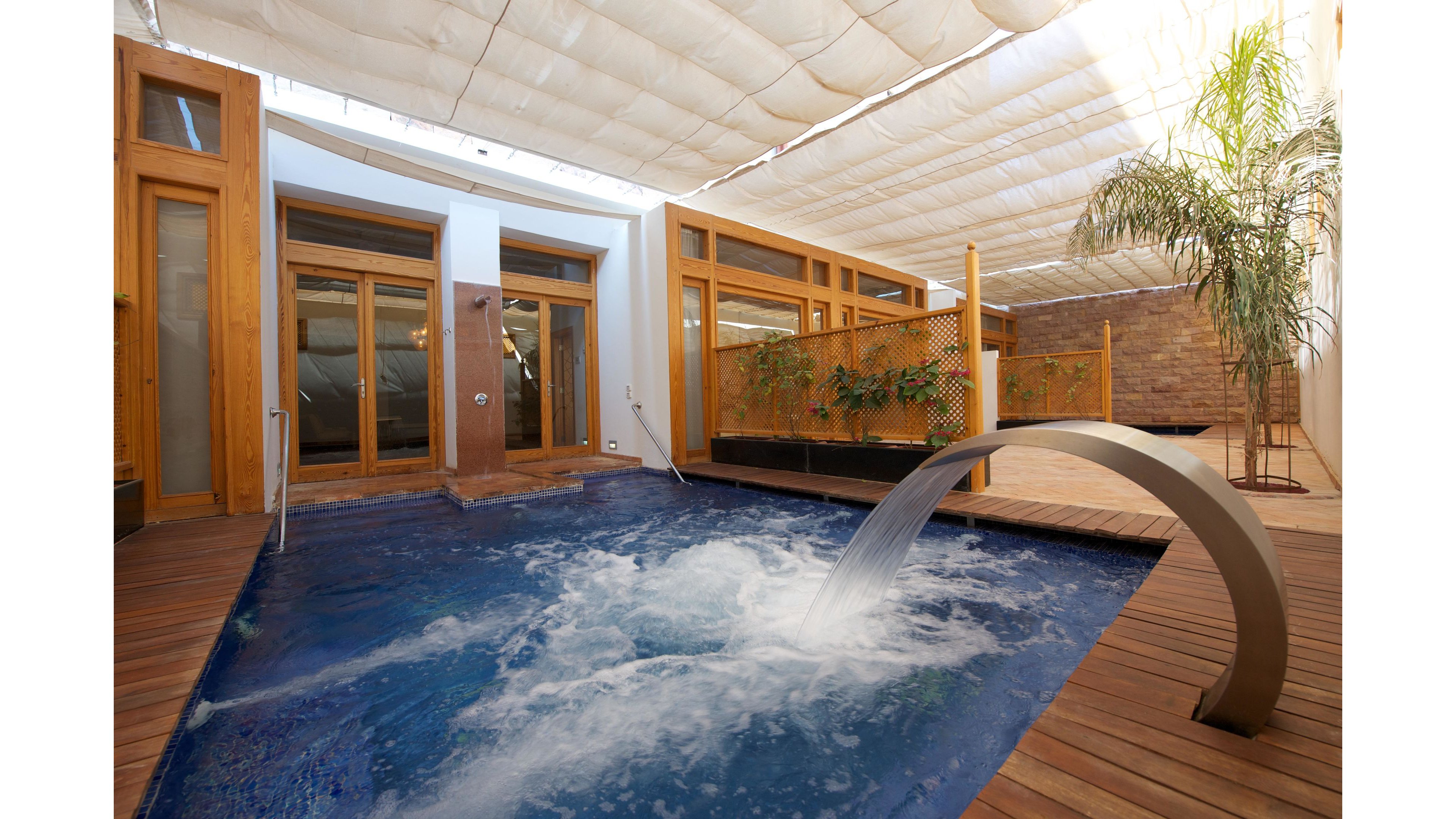 A contemporary indoor spa pool features a curved metal waterfall spout pouring into bubbling blue water. The pool is surrounded by warm wooden decking and framed by large glass doors and windows. A light fabric ceiling canopy diffuses the sunlight, creating a soft, relaxing atmosphere. Potted greenery and a tall palm tree add a touch of nature to the modern spa setting.