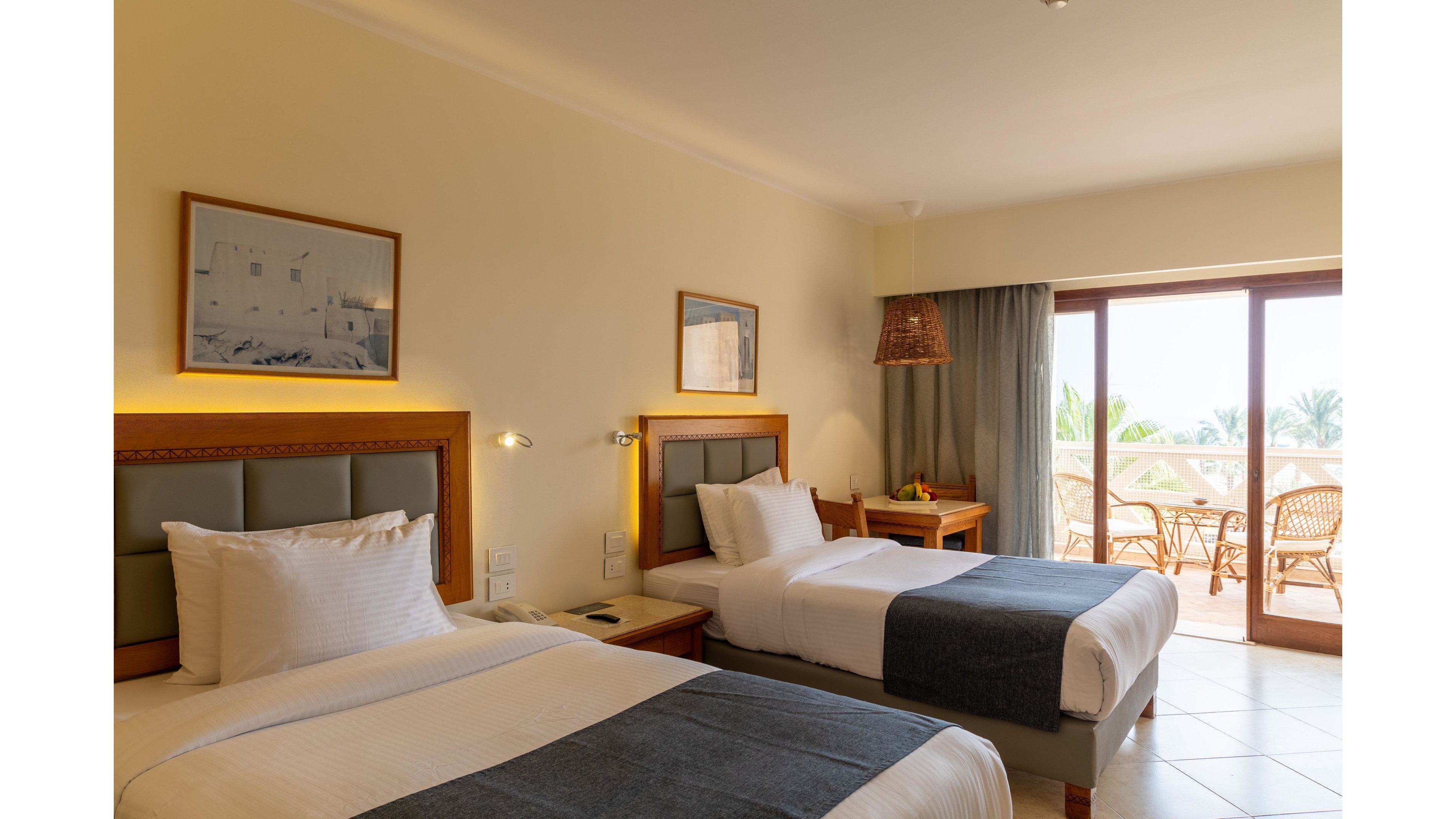 A cozy hotel room features two neatly made twin beds with white linens and dark gray throws. Warm wall lighting and framed artwork create a welcoming atmosphere against soft cream-colored walls. A sliding glass door opens to a sunlit balcony with wicker chairs and a table, overlooking palm trees and a clear sky. The overall scene feels clean, comfortable, and ideal for a relaxing vacation stay.