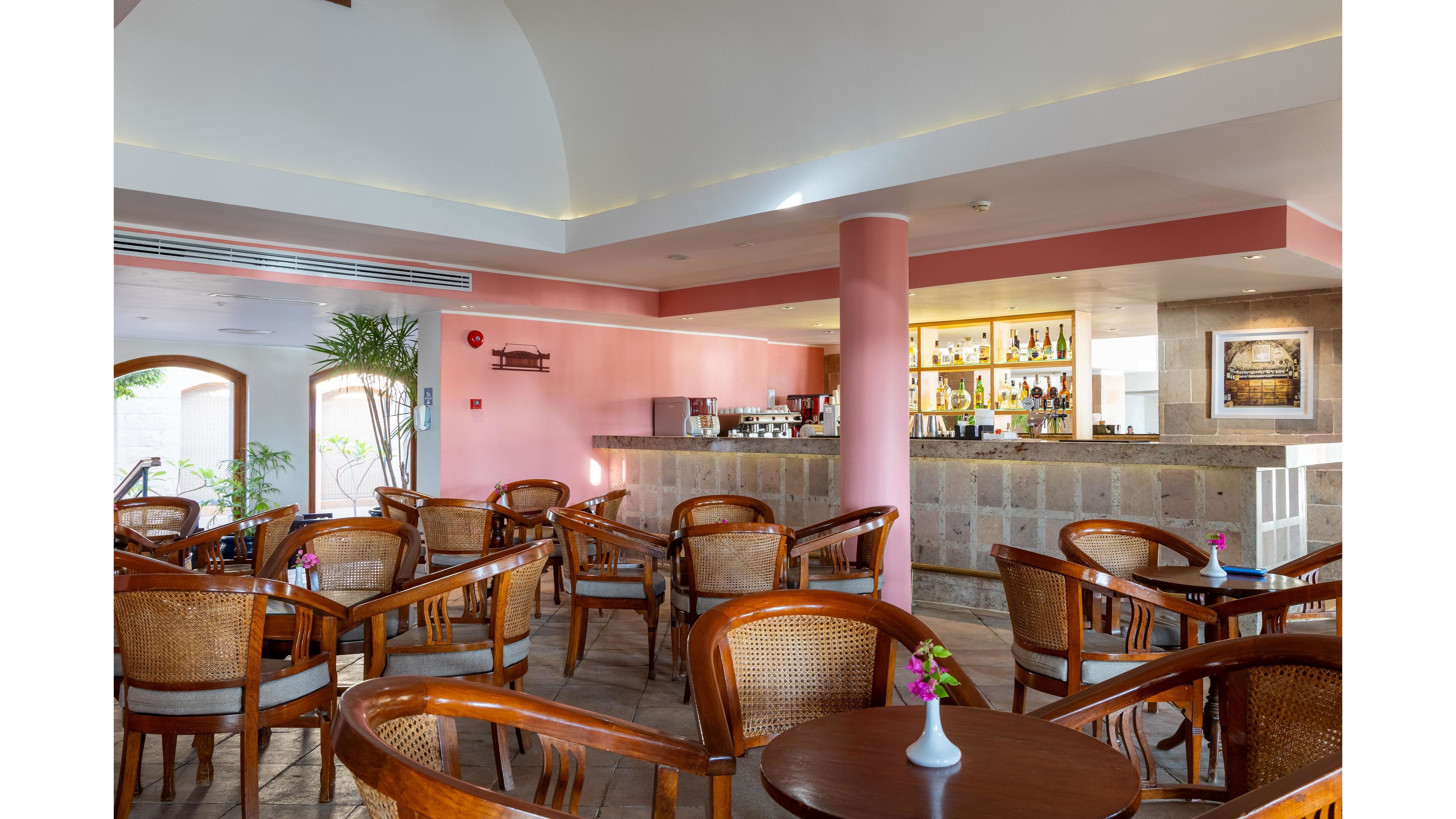 A sunlit hotel lounge bar features wooden tables and wicker armchairs arranged neatly across the tiled floor. The bar counter with stone cladding and a pink column stands in the background, displaying bottles and glassware on illuminated shelves. Large arched windows and indoor plants add natural light and greenery to the space. Small vases with colorful flowers decorate each table, creating a welcoming and relaxed atmosphere.