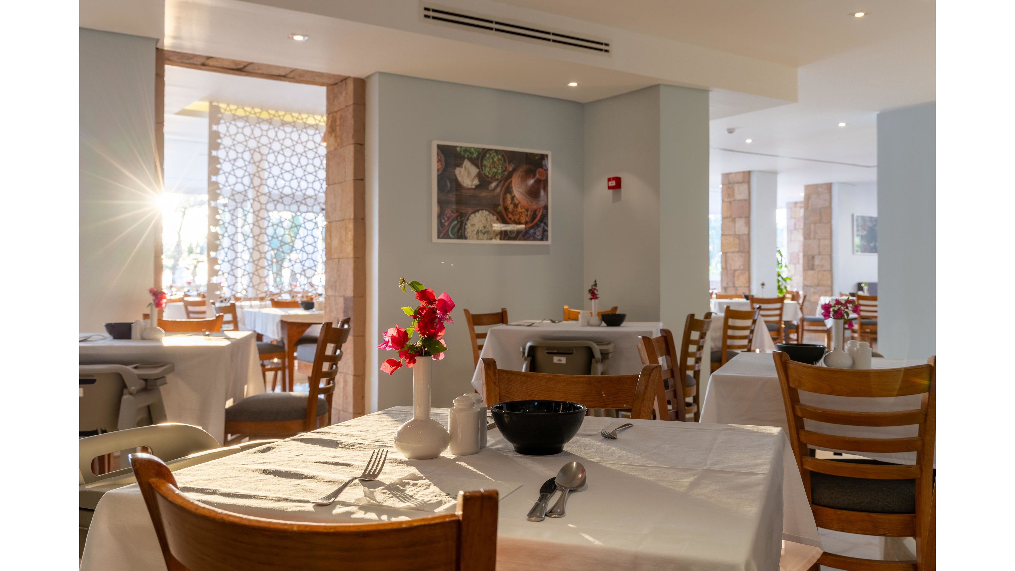 A bright restaurant dining room is shown with neatly arranged tables covered in white tablecloths. Wooden chairs surround the tables, which feature simple place settings, a black bowl, and cutlery. A small vase with vibrant pink flowers adds color to the foreground table. Sunlight streams through large windows, creating a warm and inviting atmosphere in the modern interior.
