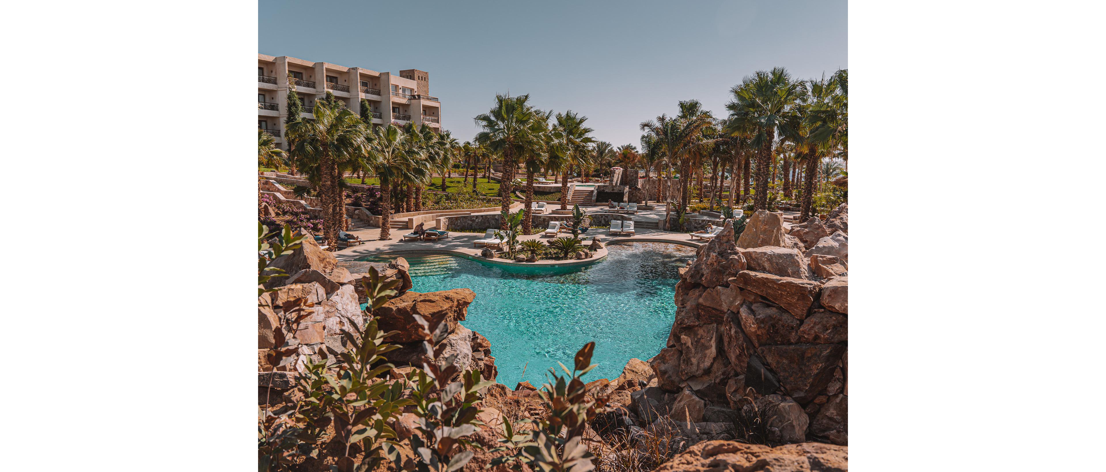 A turquoise swimming pool is nestled among natural rock formations and lush palm trees at a luxury resort. Lounge chairs and umbrellas line the poolside, suggesting a relaxing vacation atmosphere. In the background, a multi-story hotel building overlooks the landscaped grounds. The scene is bathed in warm sunlight under a clear blue sky, creating a tranquil and inviting setting.