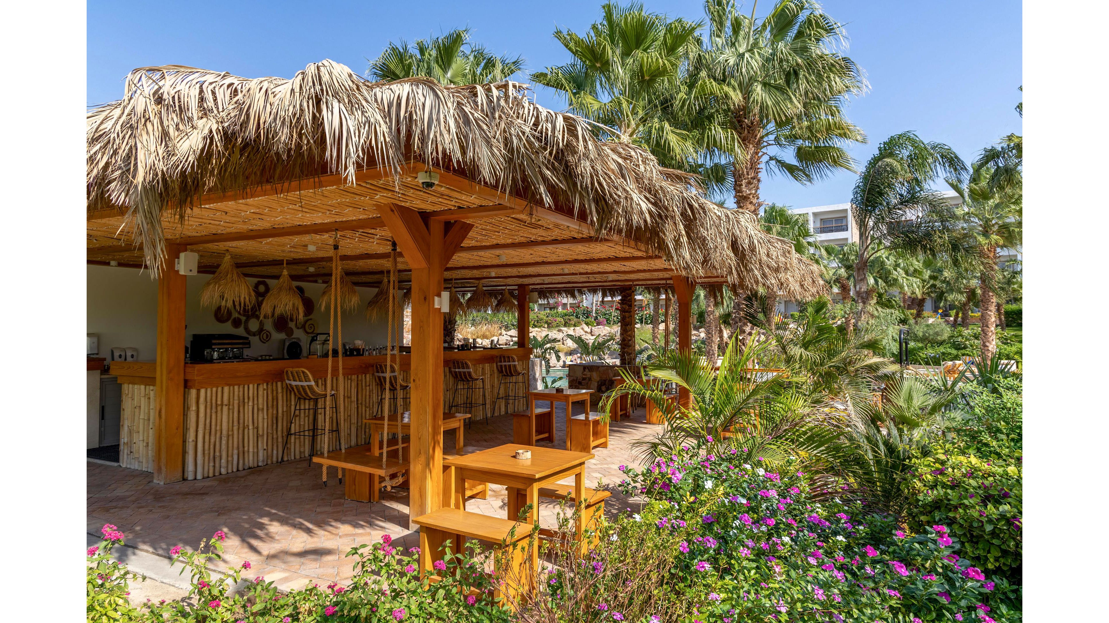 An open-air bar with a thatched roof and wooden structure sits in a lush tropical garden. Wooden tables, chairs, and swing-style bar seats are arranged under the shaded canopy. Palm trees and vibrant flowering plants surround the bar, creating a resort-like atmosphere. Bright daylight and clear blue sky enhance the relaxed vacation setting.