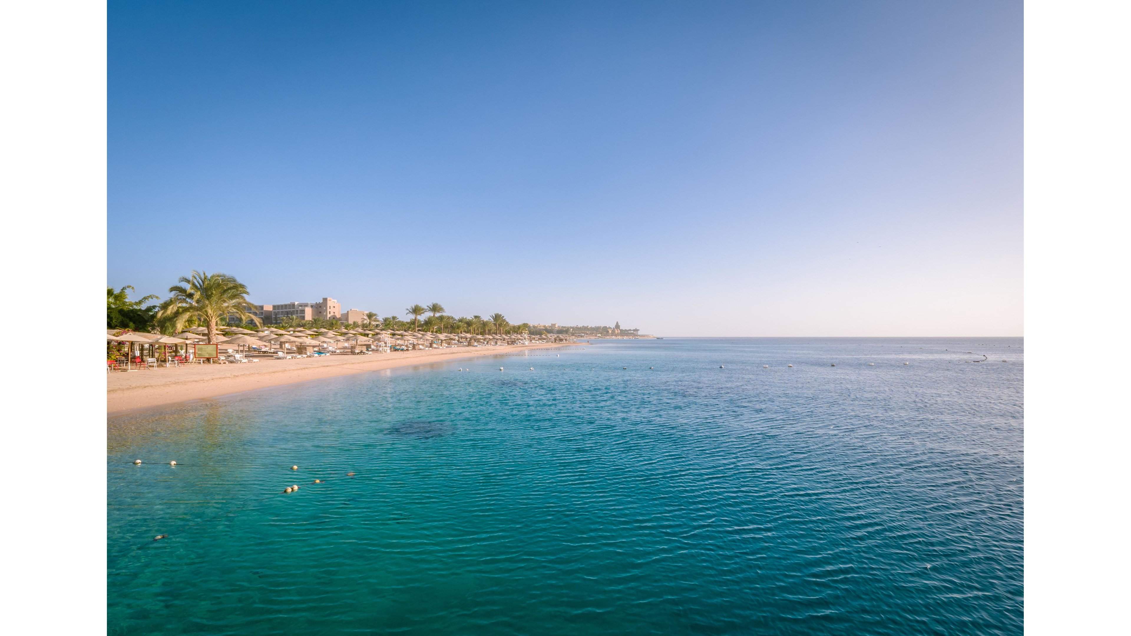 A wide sandy beach lined with sun loungers and umbrellas stretches along a calm turquoise sea. Resort buildings and palm trees are visible in the background under a clear blue sky. The water is smooth with gentle ripples, creating a peaceful coastal scene. The overall atmosphere is relaxed and inviting, suggesting a warm vacation destination.