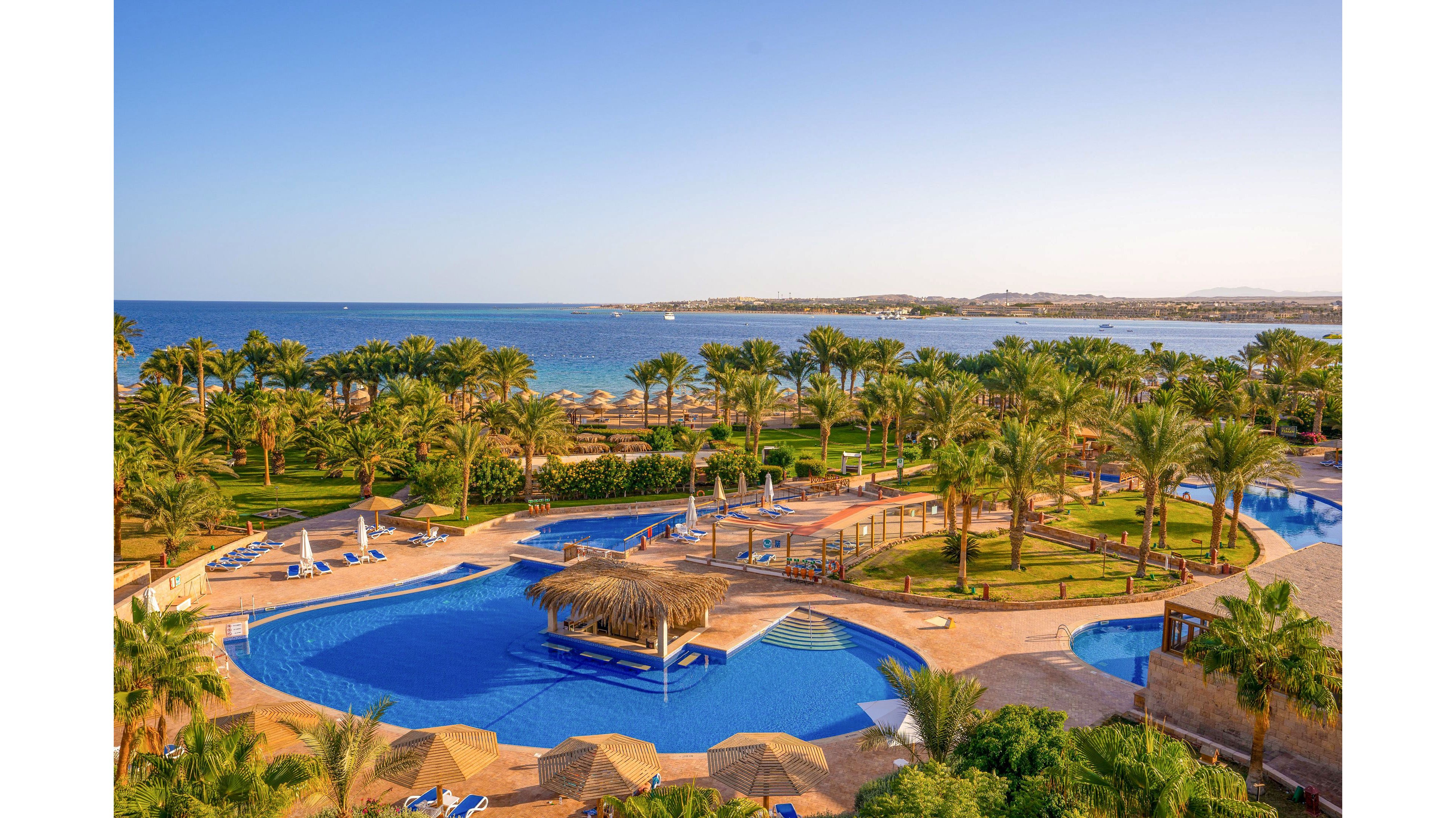 A large resort swimming pool is surrounded by palm trees and sun loungers, creating a tropical vacation atmosphere. The scene overlooks a calm blue sea and distant coastline under a clear sky. Curved pools, thatched shade structures, and landscaped lawns add to the relaxing holiday setting. The image conveys a sunny, warm climate ideal for leisure and outdoor recreation.