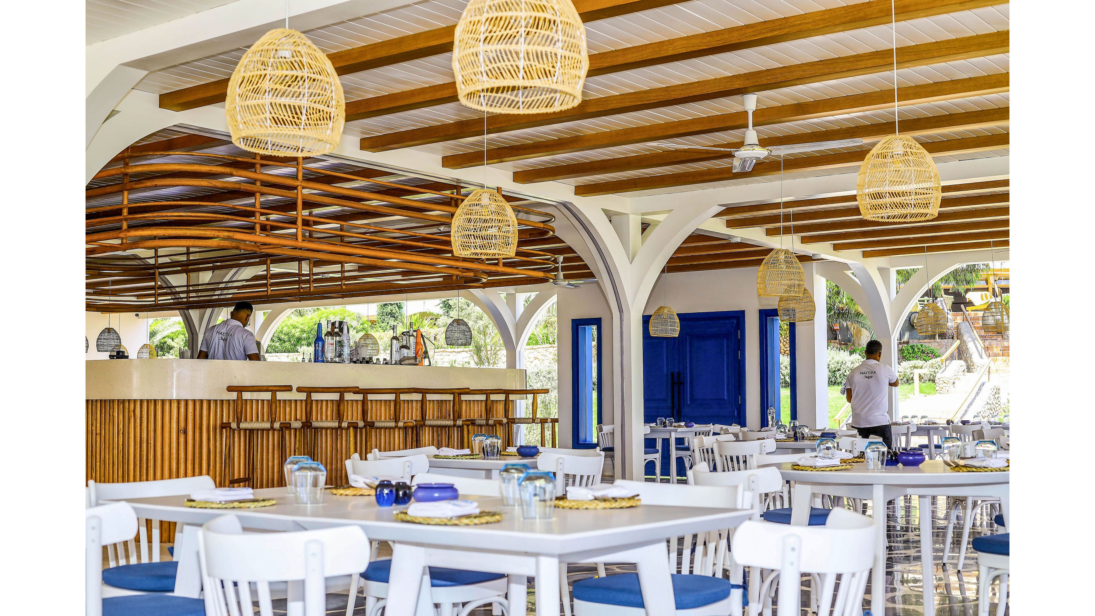 Open-air restaurant interior featuring white tables and blue cushioned chairs under a wooden beamed ceiling. Wicker pendant lamps hang above the seating area, creating a warm and inviting coastal atmosphere. A bar counter with wooden slats is visible in the background, where staff members are preparing the space. Large blue doors and windows add vibrant color accents to the bright, airy setting.