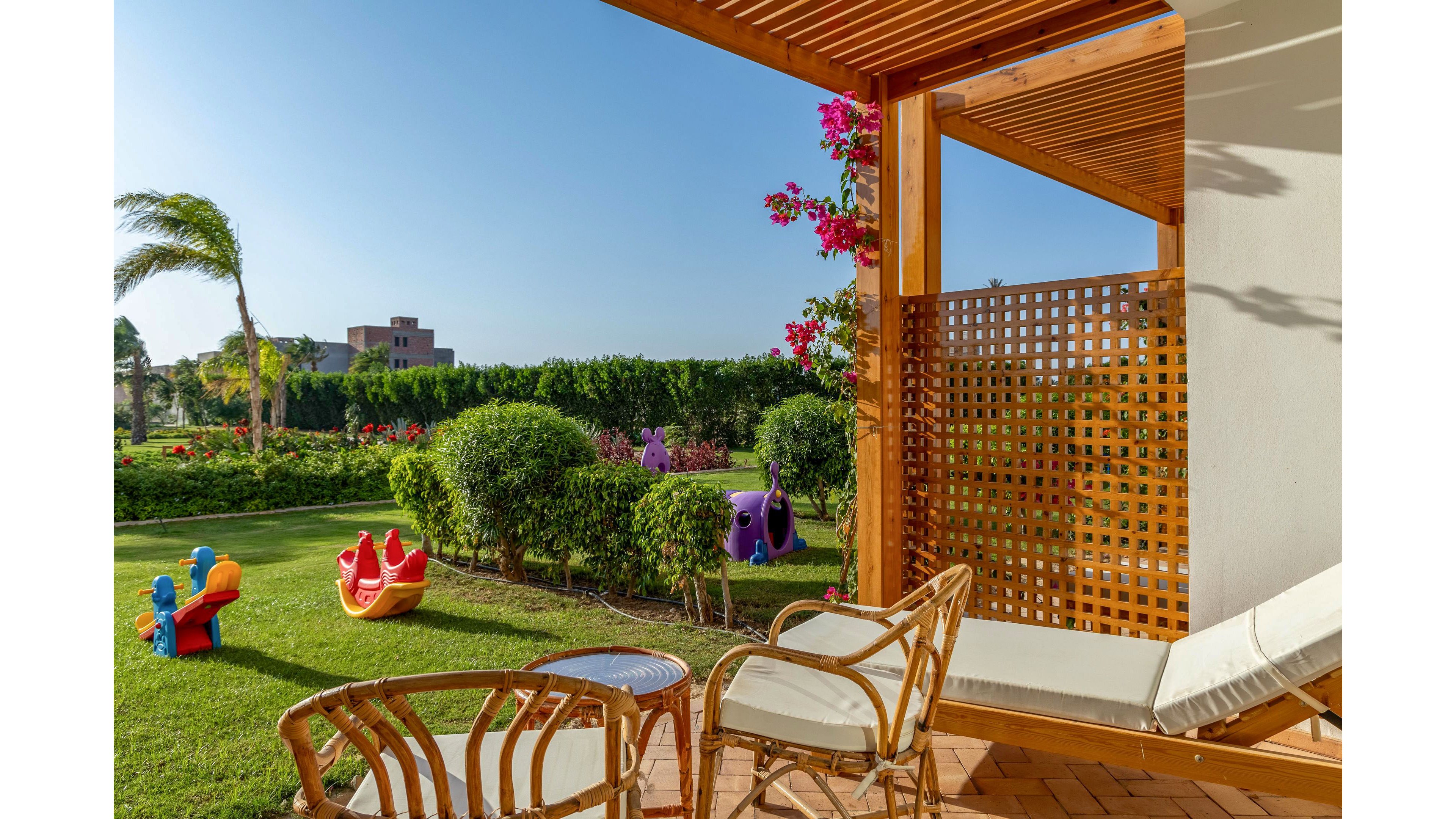 A cozy wooden terrace with wicker chairs and a lounge chair overlooks a lush green garden. Colorful children's playground toys are scattered on the grass, including animal-shaped rockers. Flowering shrubs and hedges border the lawn, with a building visible in the distant background. The scene is bright and sunny, suggesting a warm, relaxing outdoor setting.
