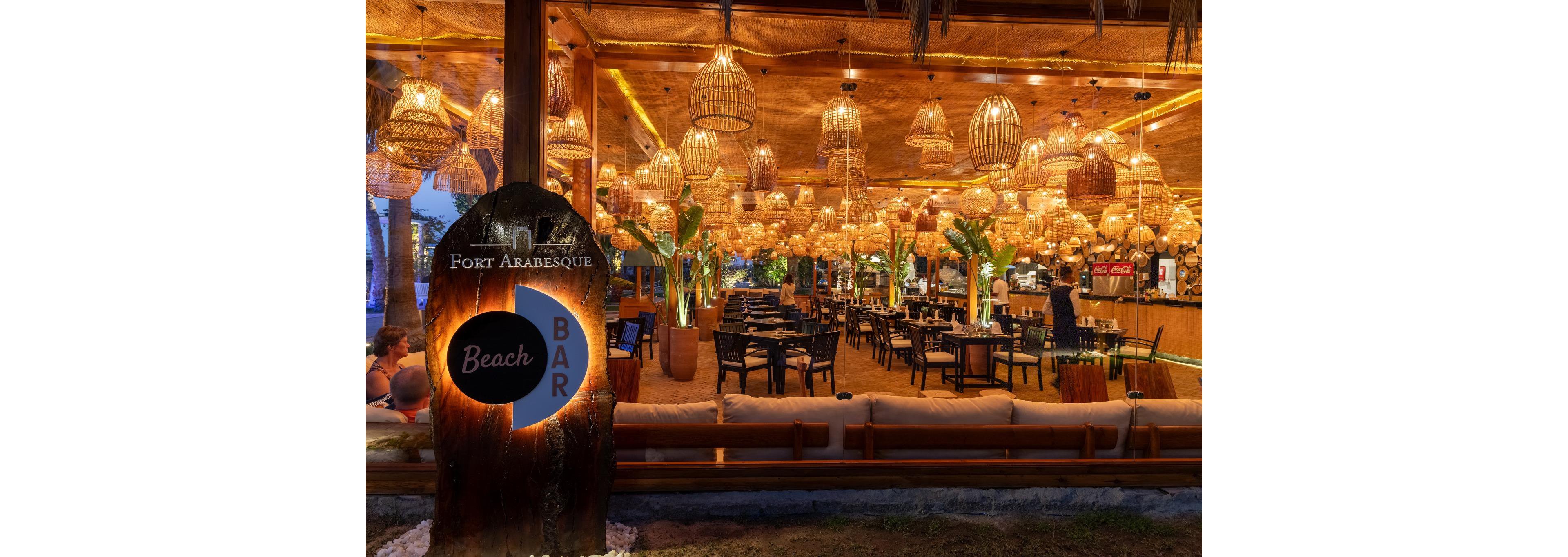 Illuminated wicker lanterns fill the Fort Arabesque Beach Bar with a warm golden glow at dusk. The open-air bar features wooden furniture, lush potted plants, and a relaxed beachfront setting. Patrons sit at tables and the bar counter, enjoying drinks in a cozy tropical atmosphere. A large wooden sign in the foreground reads "Fort Arabesque Beach Bar," welcoming guests to the venue.