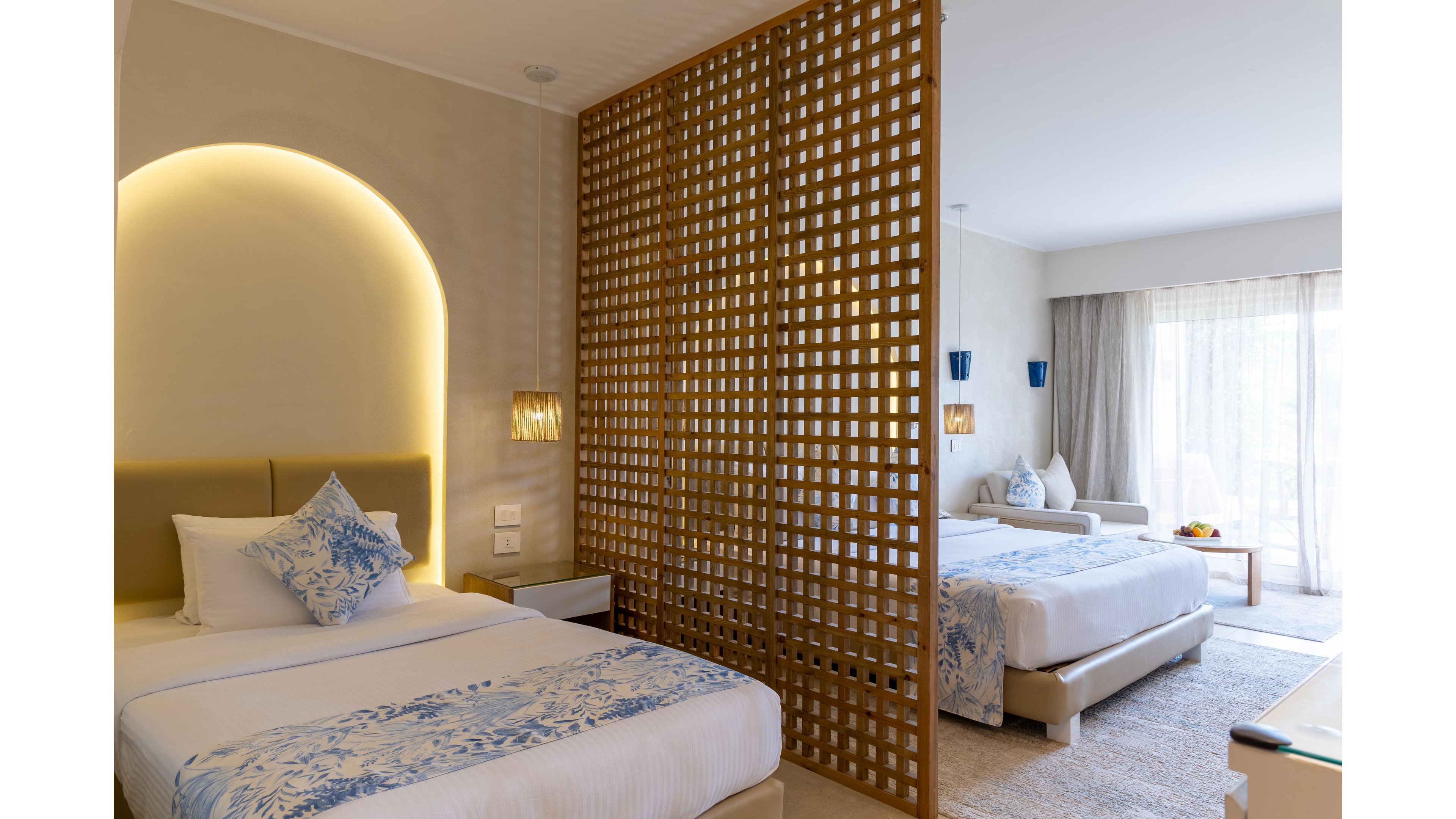 Contemporary hotel room interior featuring two neatly made twin beds separated by a decorative wooden lattice partition. Soft neutral tones and blue patterned pillows create a calm, coastal-inspired atmosphere. Warm accent lighting, wall sconces, and large floor-to-ceiling windows brighten the space. Sheer curtains, a small table with fruit, and minimal decor emphasize comfort and relaxation.