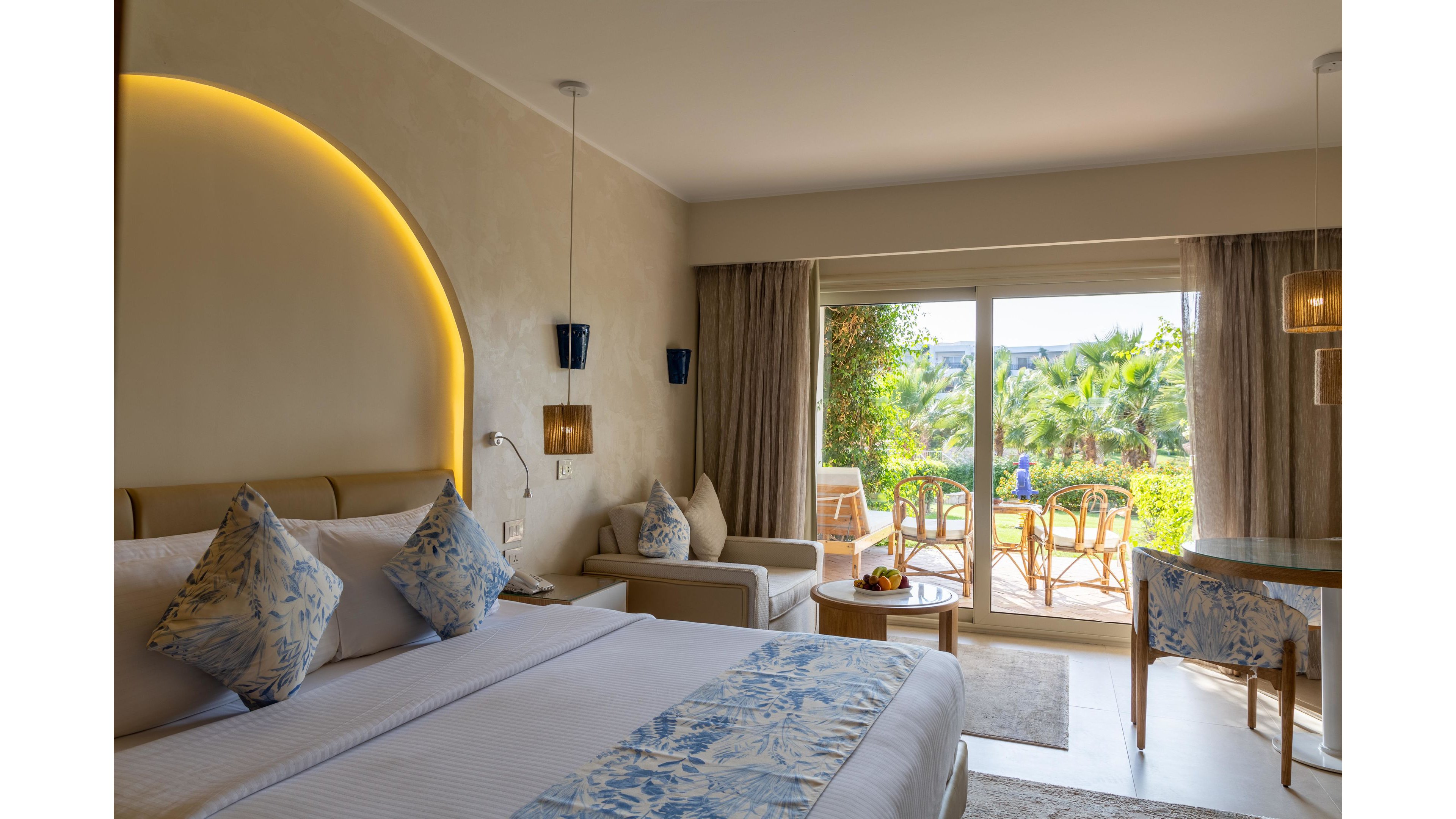 A contemporary hotel room features a large bed with patterned blue and white pillows and bedding. Soft warm lighting and neutral walls create a cozy, inviting atmosphere, complemented by a small sofa and side table. Floor-to-ceiling glass doors open onto a terrace with chairs and a table, overlooking lush green palm trees and garden scenery. The overall setting suggests a relaxing vacation stay in a resort environment.