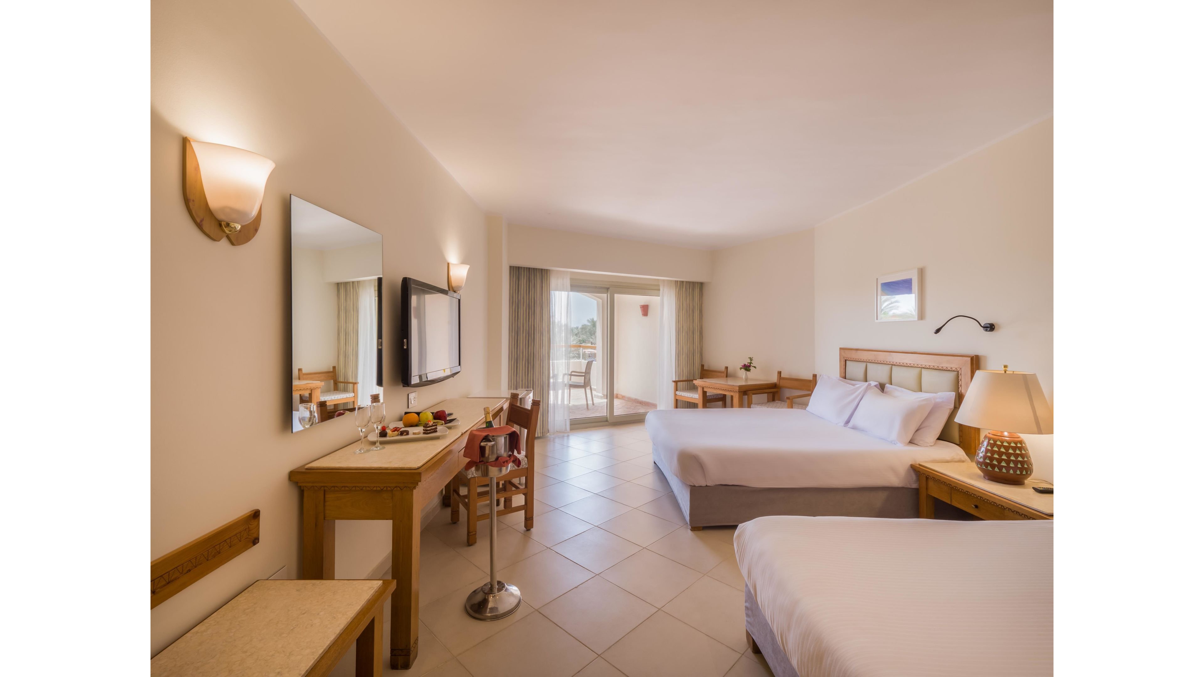 Spacious hotel room features two neatly made beds and light-colored tiled flooring. A wooden desk with a mirror, television, and fruit tray sits along one wall, while a small dining table is placed near the balcony doors. Large sliding glass doors open to a balcony with outdoor chairs and a scenic view. Soft lighting from wall sconces and a bedside lamp creates a warm, inviting atmosphere.