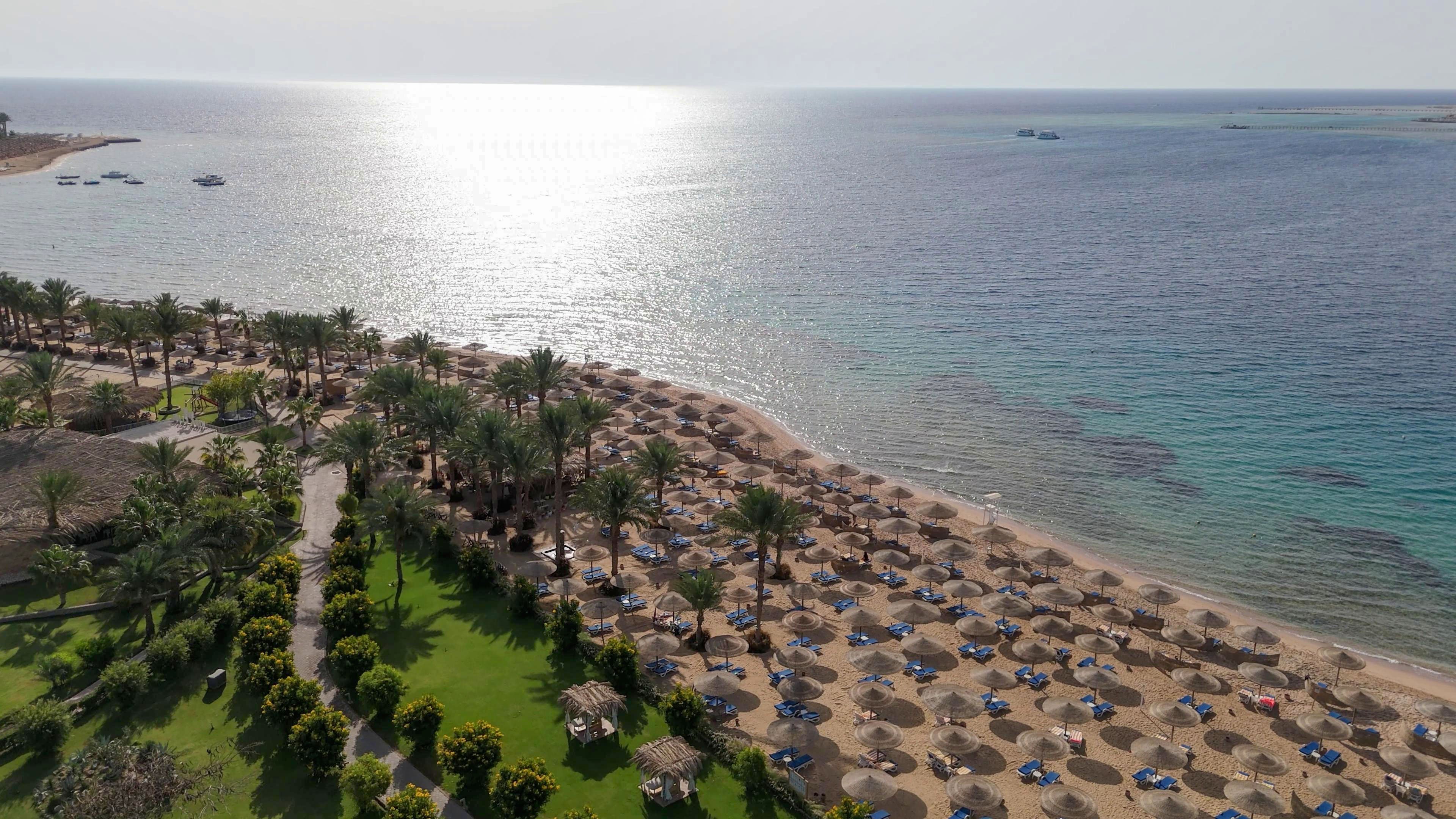 A wide aerial view shows a sunlit tropical beach resort along a calm sea. Numerous straw umbrellas and sun loungers line the sandy shoreline, facing the shimmering water. Lush green lawns and palm trees border the beach, with small huts and pathways weaving through the landscaped area. The overall scene suggests a peaceful vacation destination with clear weather and gentle waves.