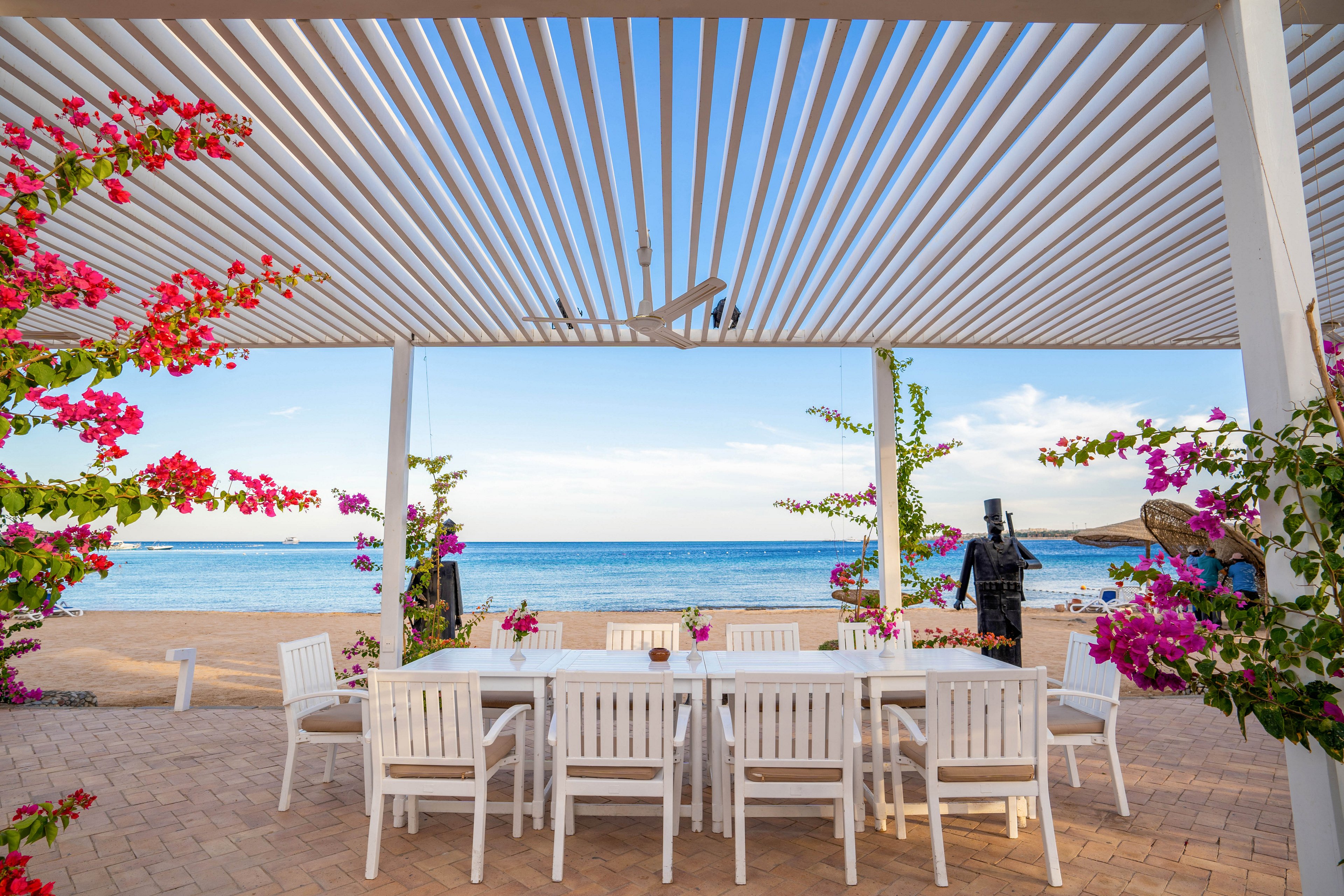 A spacious outdoor dining area features a long white table and matching chairs under a modern white pergola. The setting overlooks a tranquil blue sea, with vibrant pink and magenta flowers climbing the pergola structure. The scene is bright and inviting, evoking a relaxed, summery mood with clear skies and coastal ambiance.