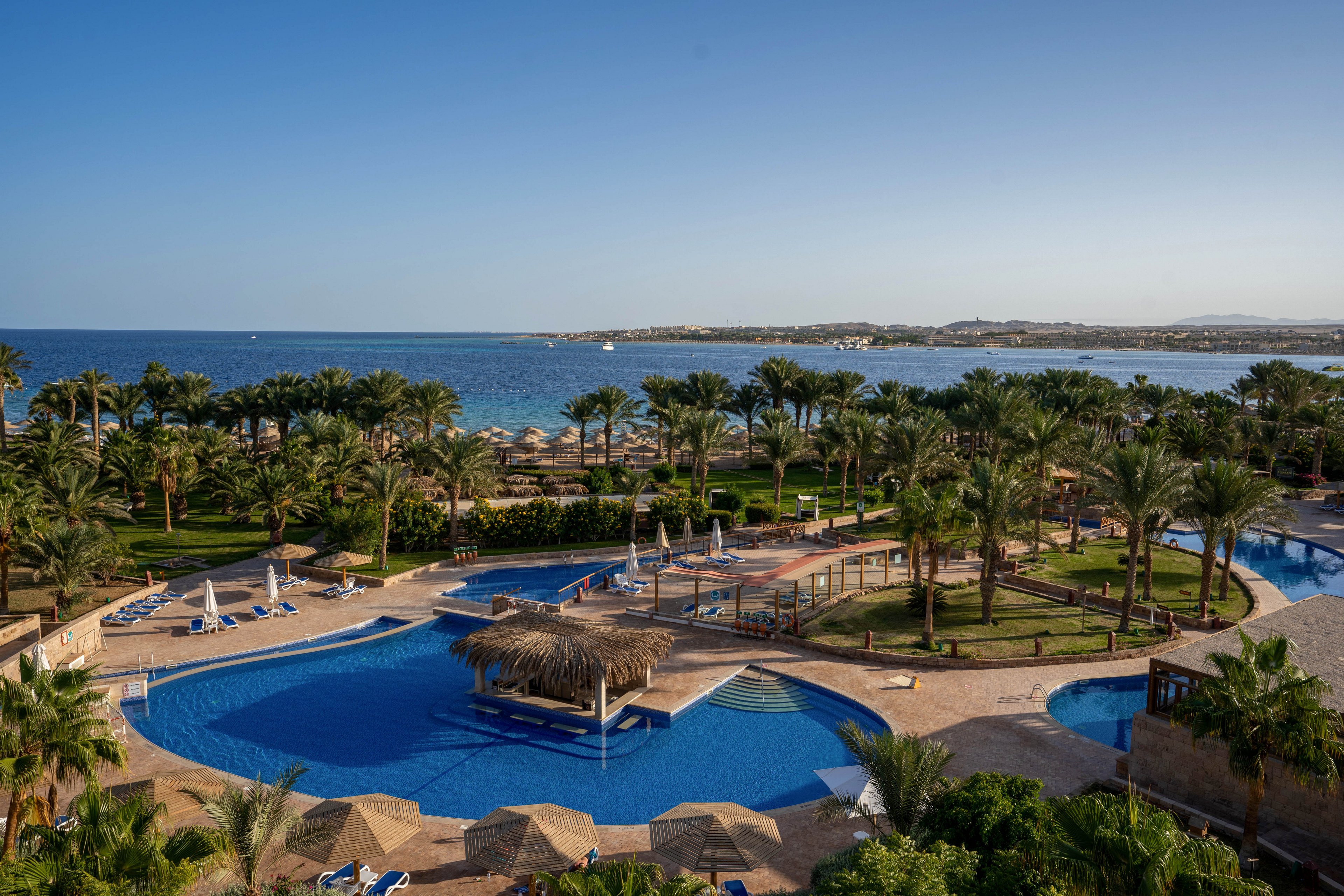 A luxurious resort features a large, curving swimming pool surrounded by palm trees and sun loungers. The setting is tropical, with clear blue skies and a calm sea visible in the background. Thatched umbrellas and shaded seating areas provide relaxation spots for guests. The overall mood is tranquil and inviting, ideal for a vacation or getaway. No visible text or numbers are present in the image.